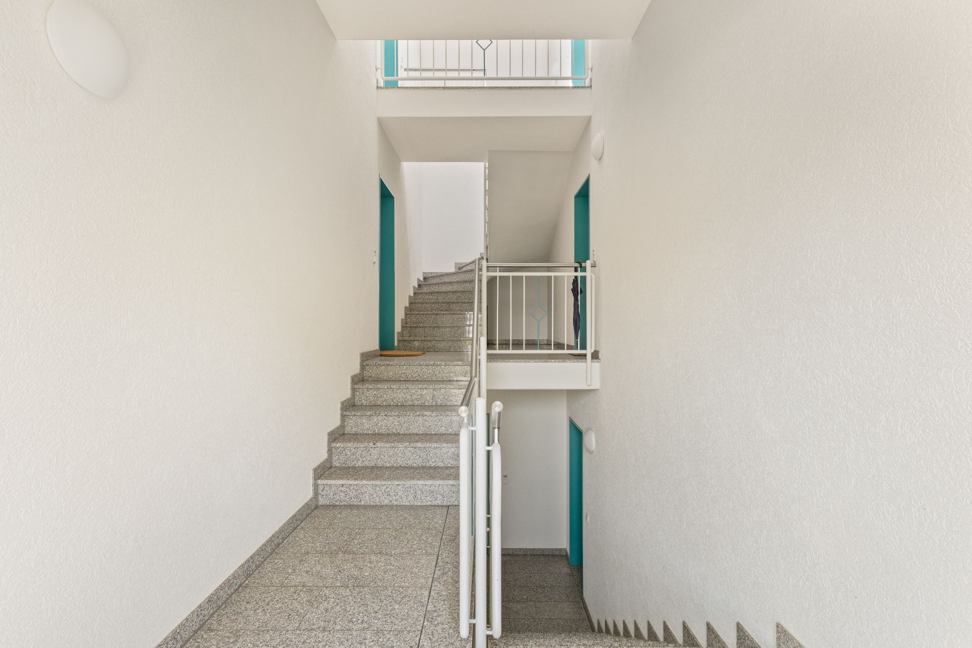 Helles Treppenhaus mit grauen Stufen, weißen Wänden und türkisfarbenen Türrahmen.