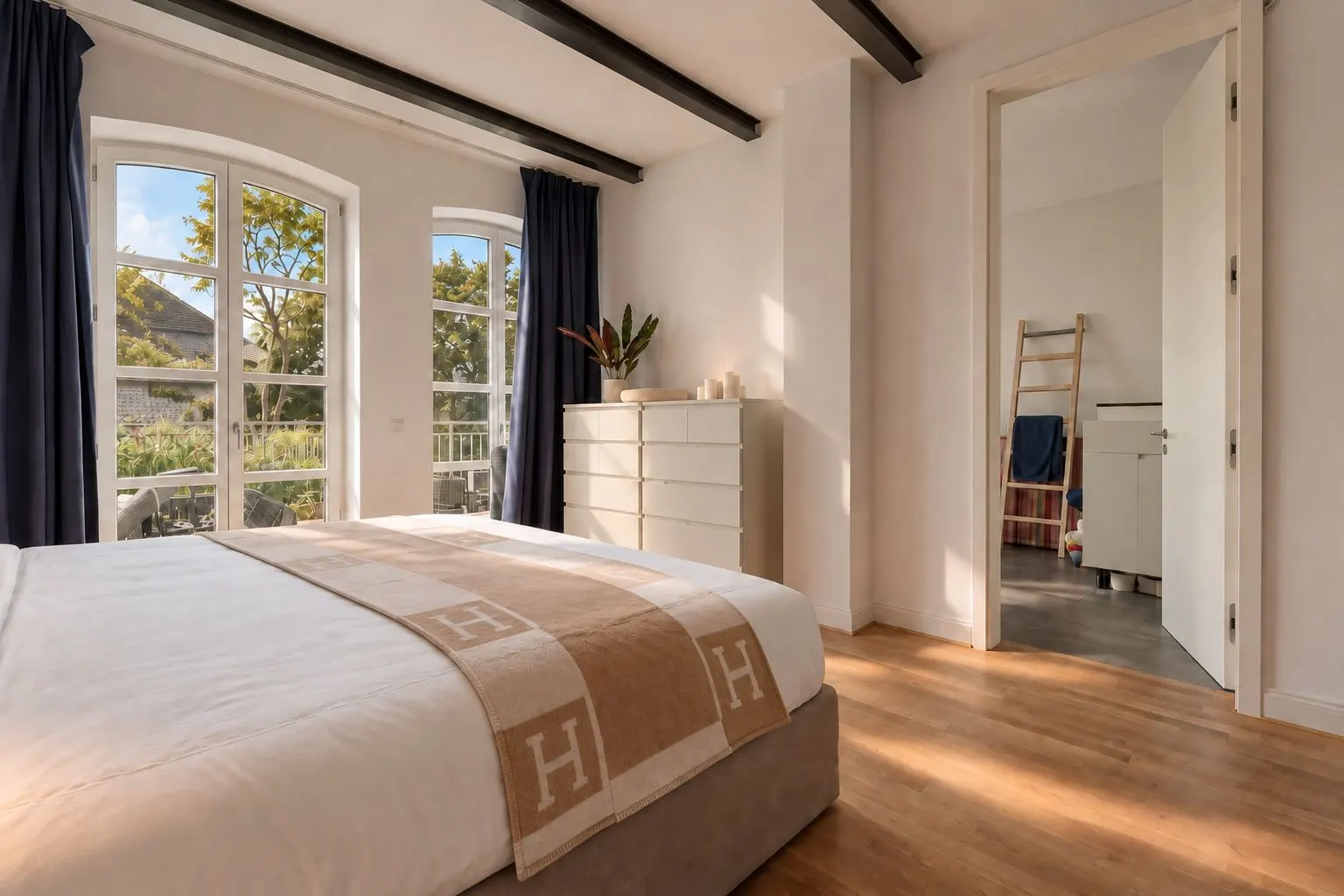 Helles Schlafzimmer mit Doppelbett, Kommode, Holzfußboden und großen Fenstern mit Blick ins Grüne.