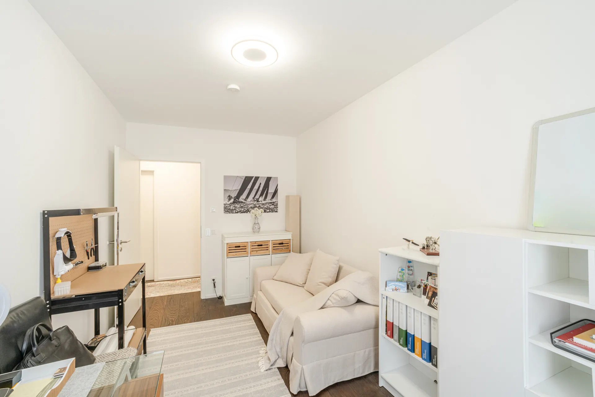 Helles Zimmer mit Sofa, Regal, Werkbank und Bild an der Wand.