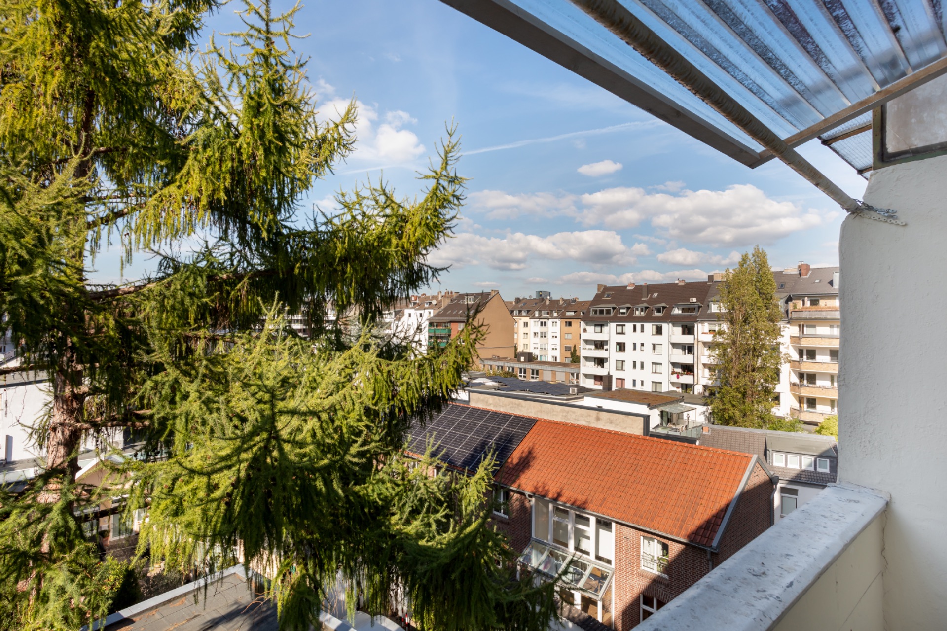 Blick von einem Balkon auf Wohngebäude, Bäume und blauen Himmel mit Wolken.