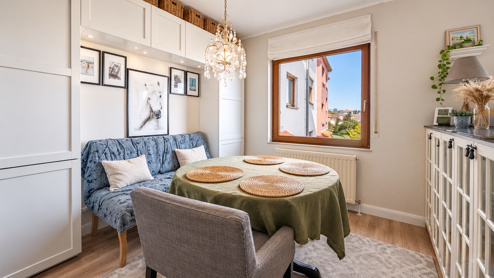 Heller Essbereich mit Sofa, rundem Tisch, Fenster und dekorativem Kronleuchter.