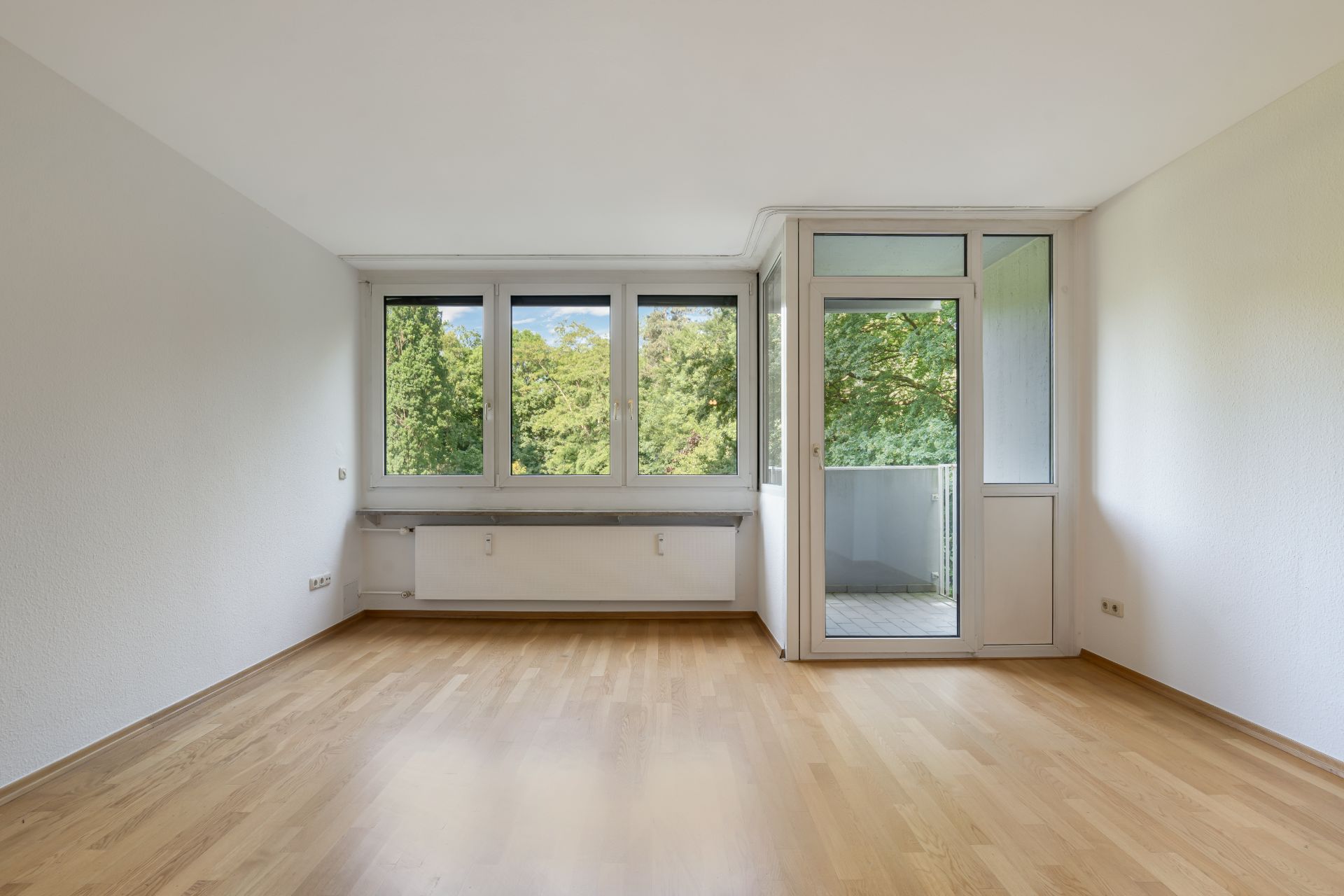 Leeres Zimmer mit Holzfußboden, großen Fenstern und Zugang zu einem Balkon.