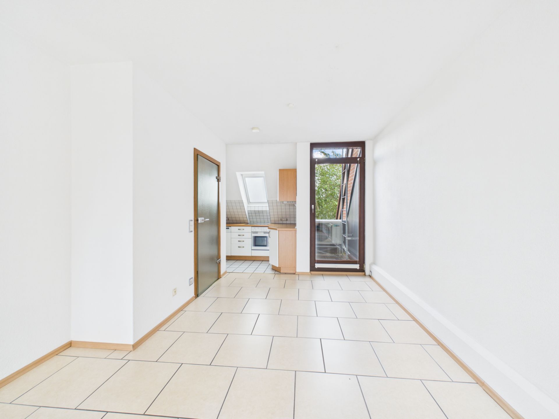 Heller Wohnraum mit gefliesten Böden, kleiner Einbauküche und Zugang zu einem Balkon.