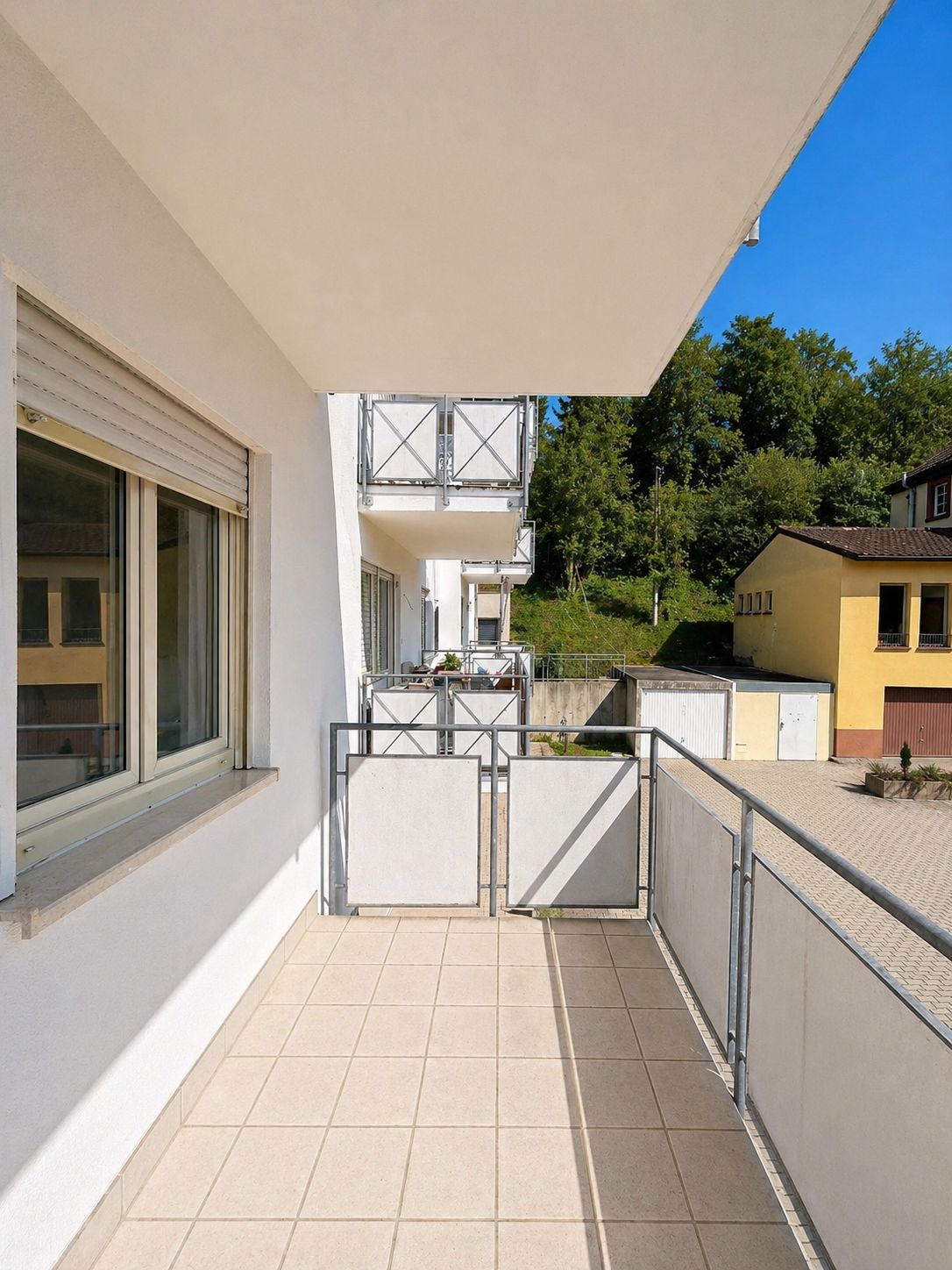Heller Balkon mit Fliesenboden, Geländer und Blick auf Nachbargebäude und Bäume.