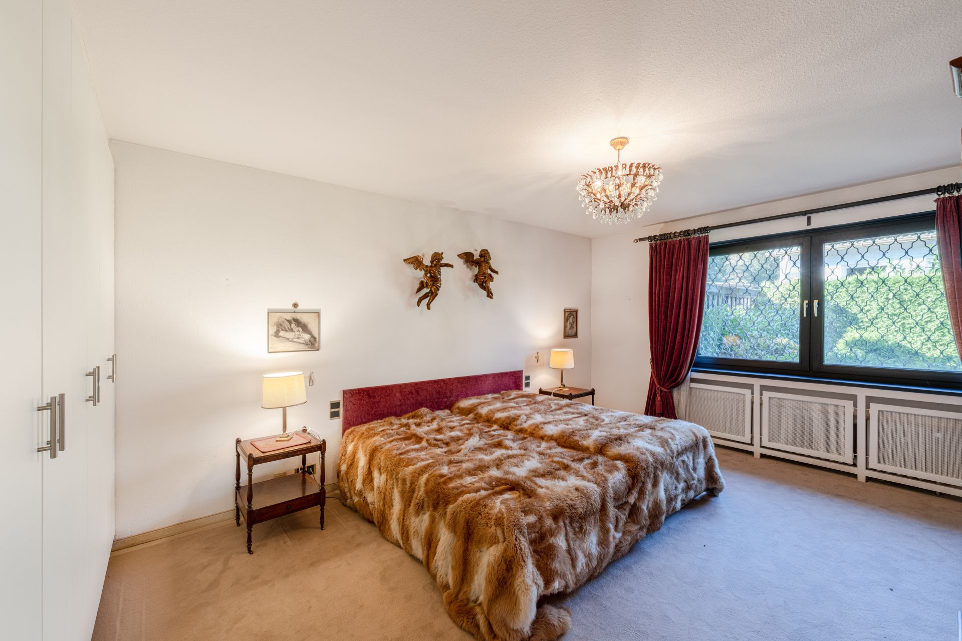 Schlafzimmer mit großem Bett, Felldecke, Kronleuchter und Fenster mit roten Vorhängen.