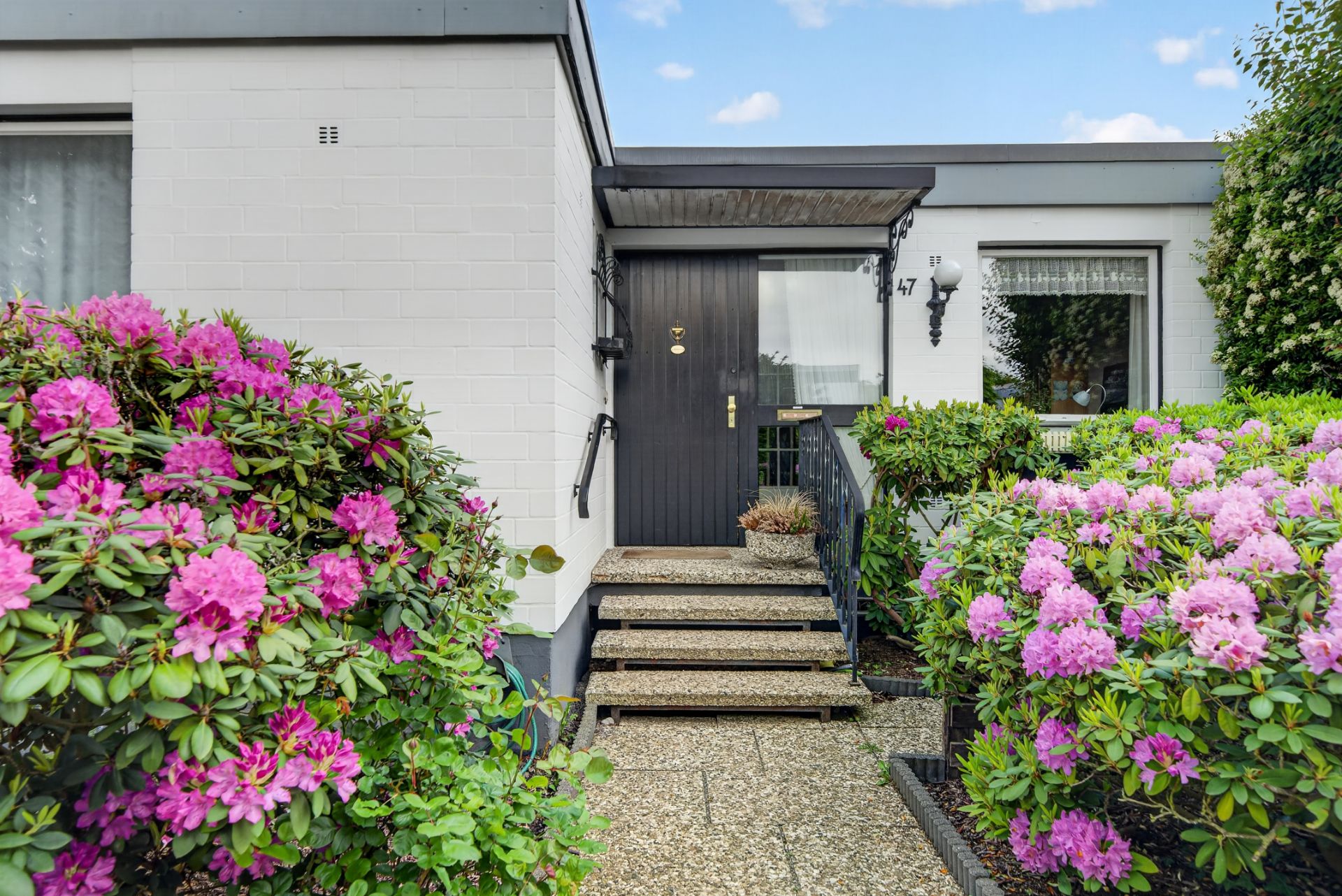 Eingang eines Hauses mit Steintreppe, blühenden Rhododendron-Büschen und schwarzer Tür.