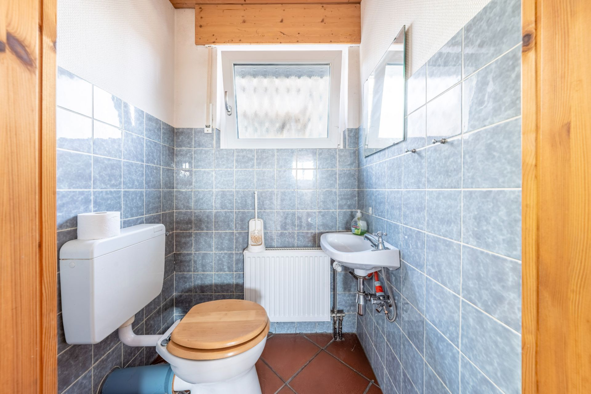 Kleines Badezimmer mit WC, Waschbecken, Spiegel und Fenster, blaue Fliesen an den Wänden.