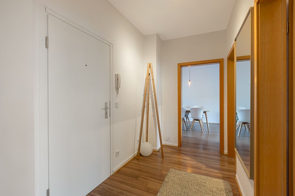 Heller Flur mit Holzboden, großem Spiegel und Blick in einen Essbereich mit weißen Stühlen.
