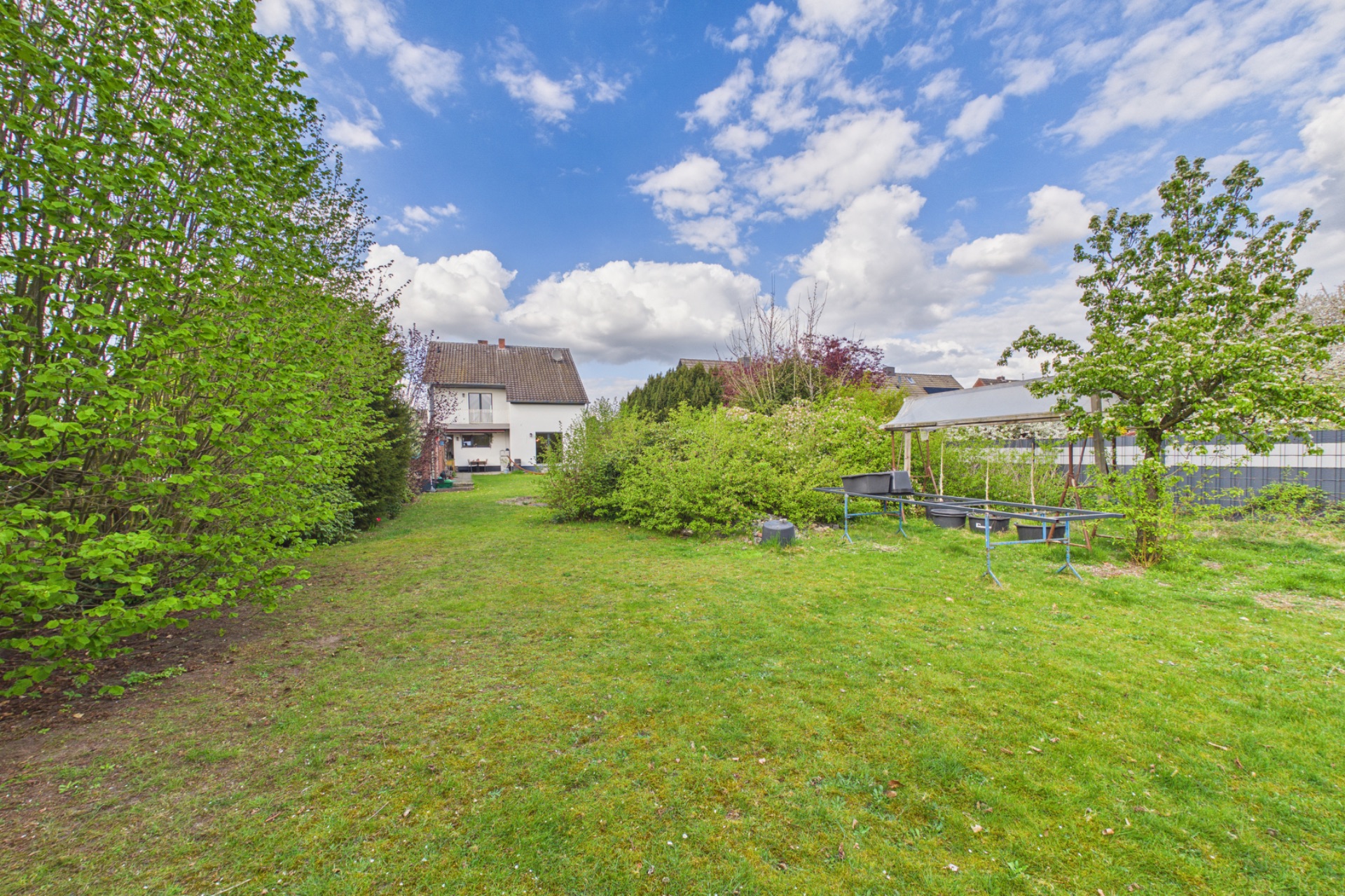 Großer grüner Garten mit Rasen, Bäumen, Sträuchern und einem weißen Wohnhaus im Hintergrund.