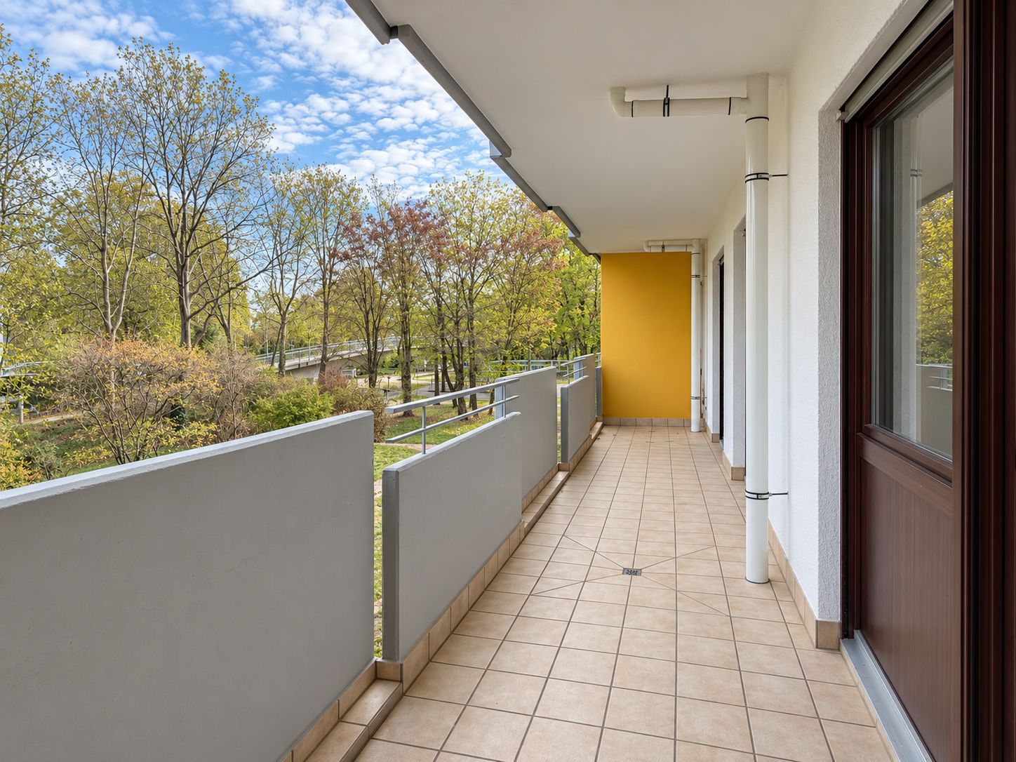 Langer, gefliester Balkon mit Geländer und Blick auf grüne Bäume.