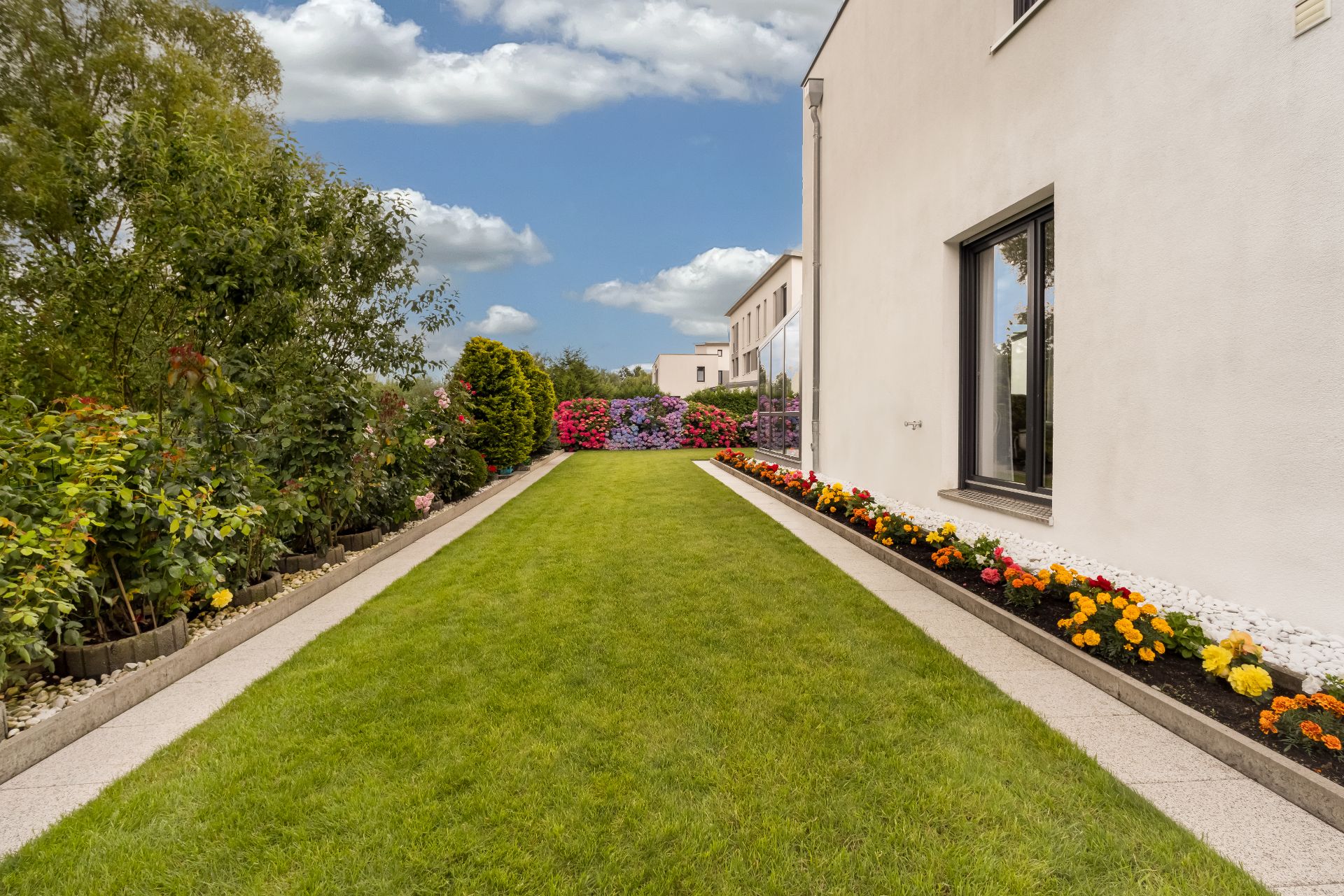 Langer Garten mit Rasen, bunten Blumenbeeten und modernen Gebäuden an der Seite.