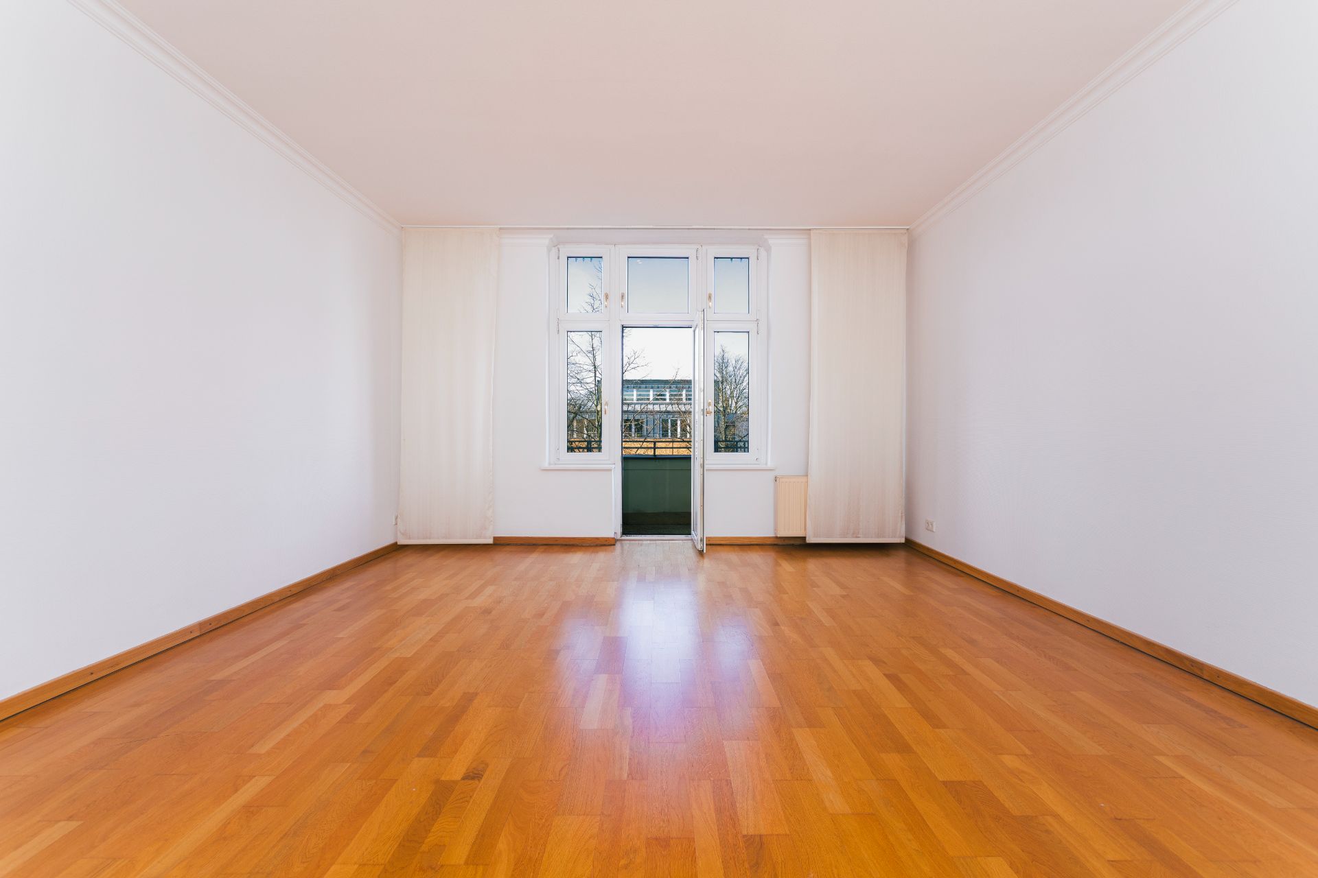 Helles, leeres Zimmer mit Holzboden und großem Fenster zur Straße.