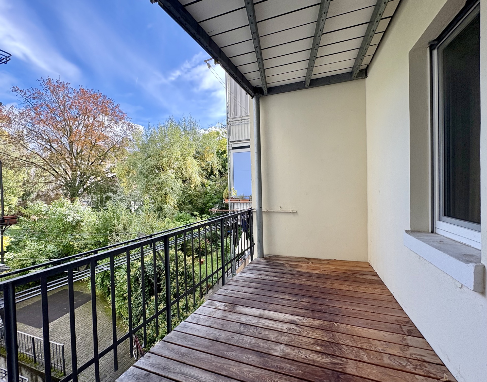 Überdachter Balkon mit Holzboden und Metallgeländer, Blick auf grüne Bäume.