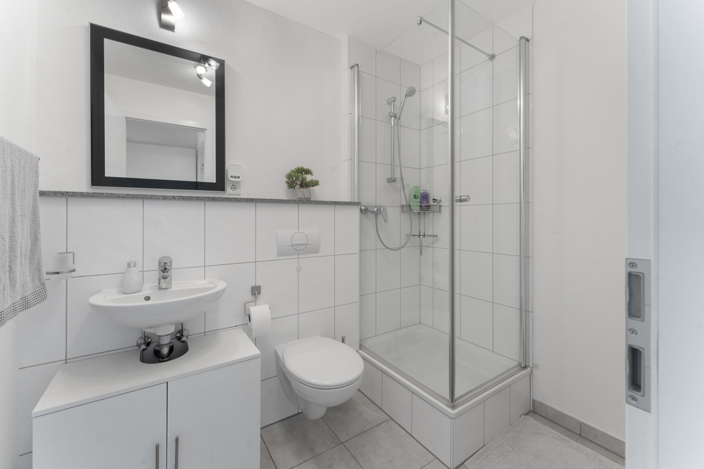 Modernes, weiß gefliestes Badezimmer mit Dusche, Waschbecken, Spiegel und Toilette.