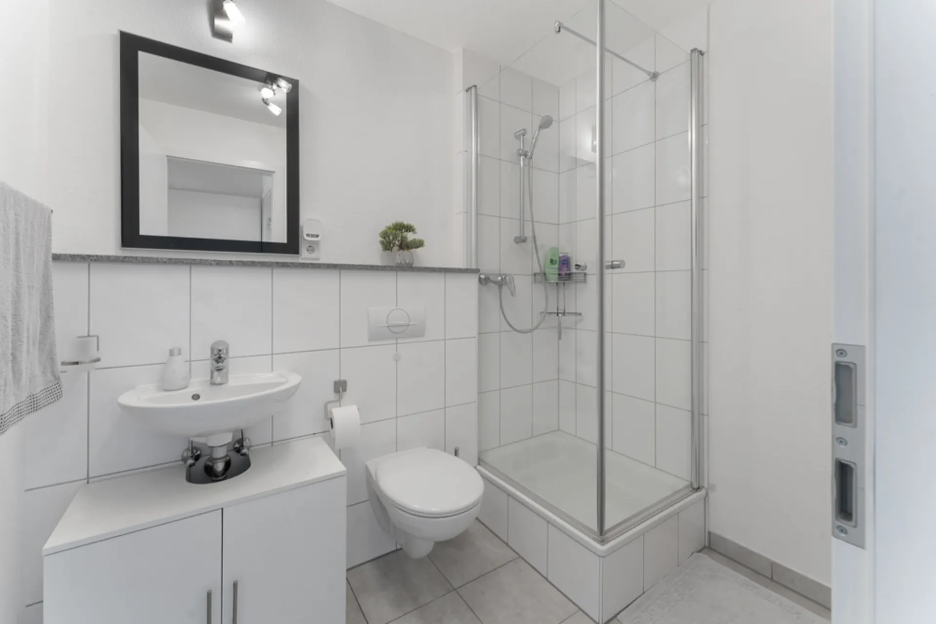 Modernes, weiß gefliestes Badezimmer mit Dusche, Waschbecken, Spiegel und Toilette.