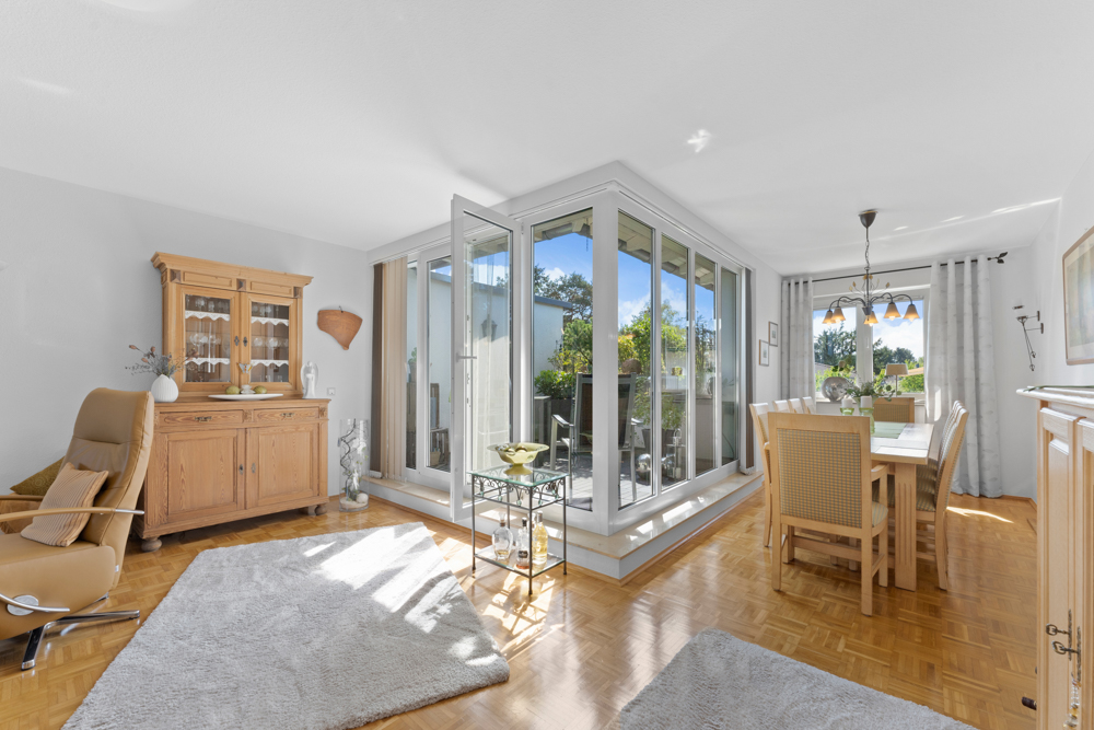 Helles Wohnzimmer mit Essbereich, Holzmöbeln, Teppich und Zugang zu einem Balkon durch große Glasfenster.