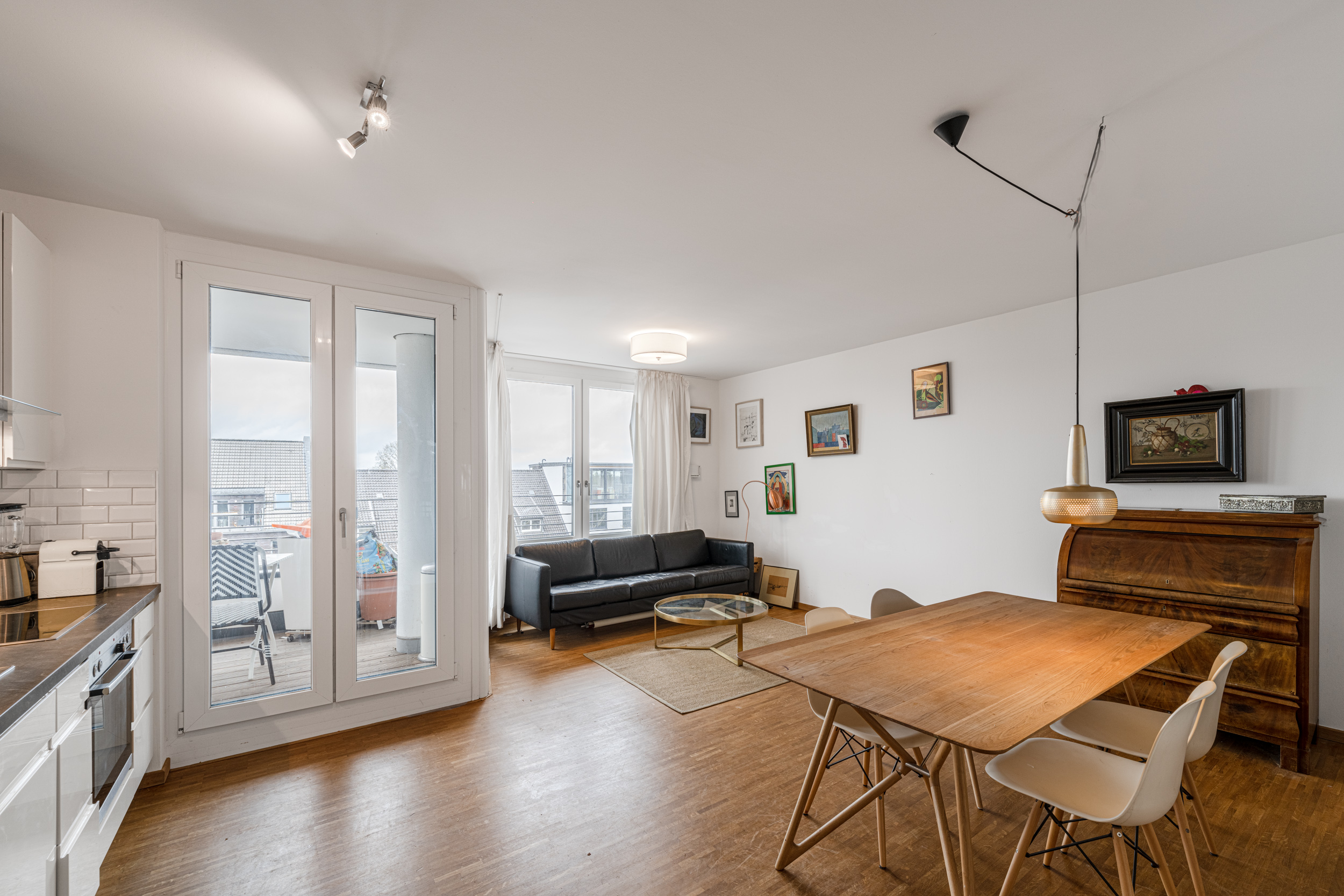 Helles Wohnzimmer mit Holzboden, Sofa, Esstisch, Küchenzeile und Zugang zu einem Balkon.