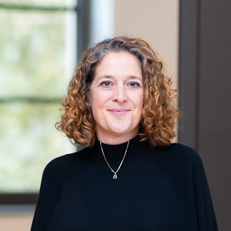 Eine Frau mit lockigen Haaren trägt einen schwarzen Pullover und steht vor einem Fenster.