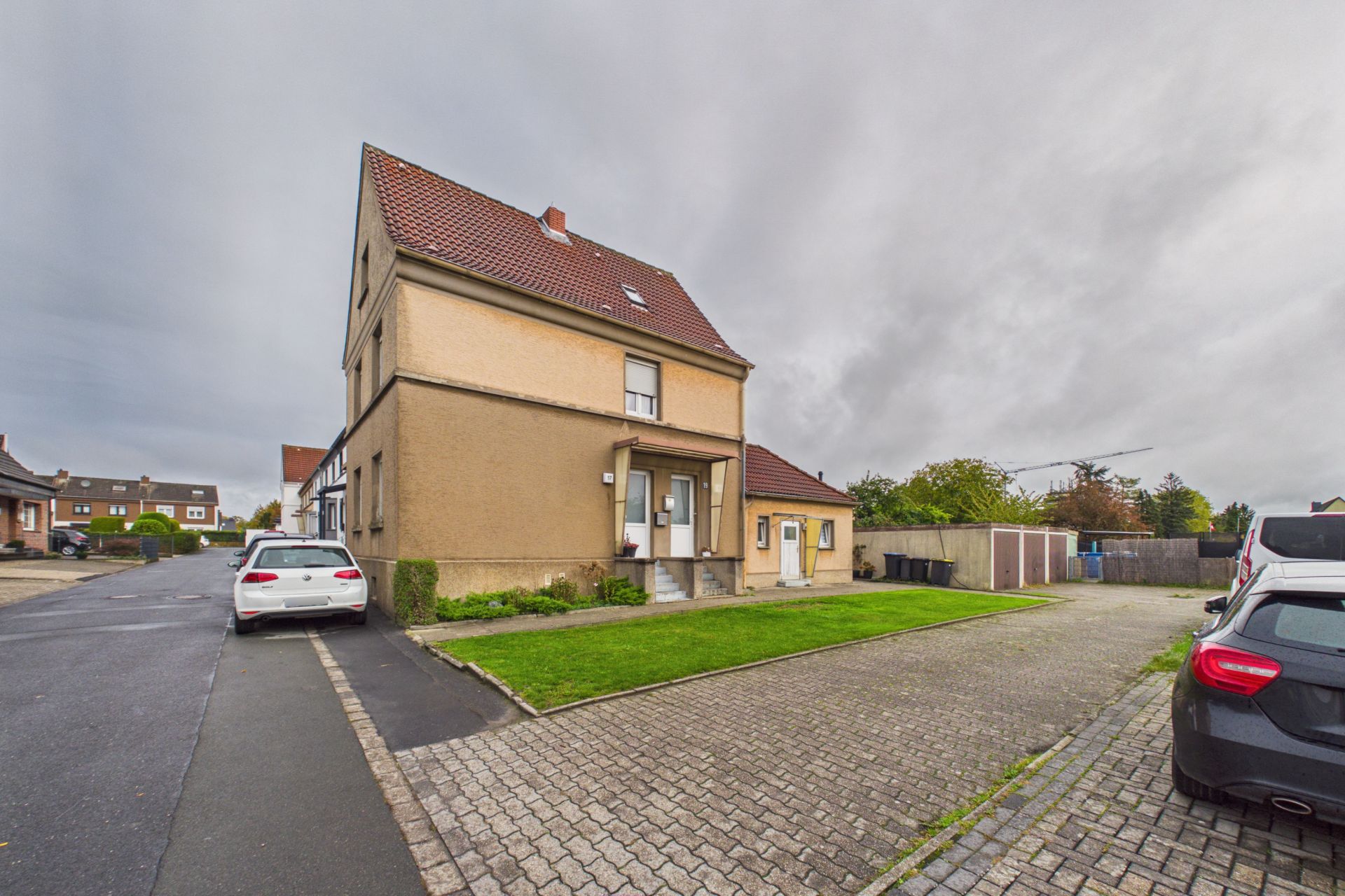 Zweistöckiges Wohnhaus mit rotem Dach, Vorgarten und parkenden Autos auf gepflasterter Einfahrt.