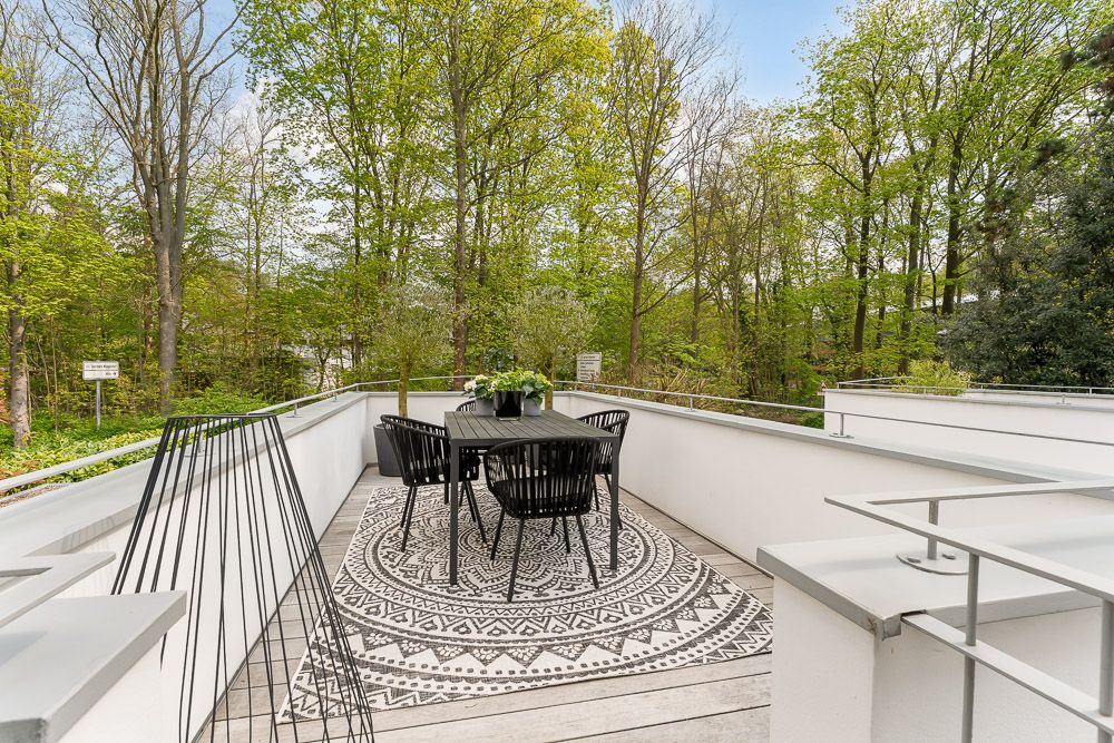Balkon mit Tisch, vier schwarzen Stühlen und Teppich, umgeben von Bäumen.