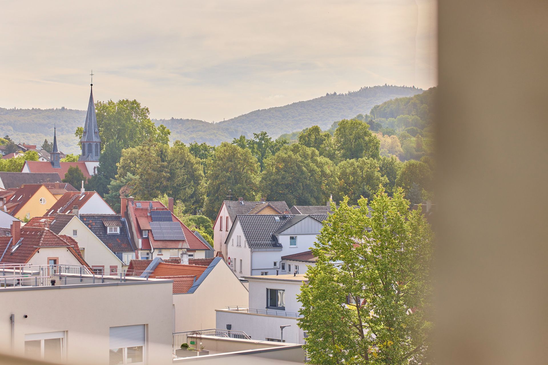 Blick auf Wohnhäuser mit roten Dächern, Bäumen und einem Kirchturm im Hintergrund.