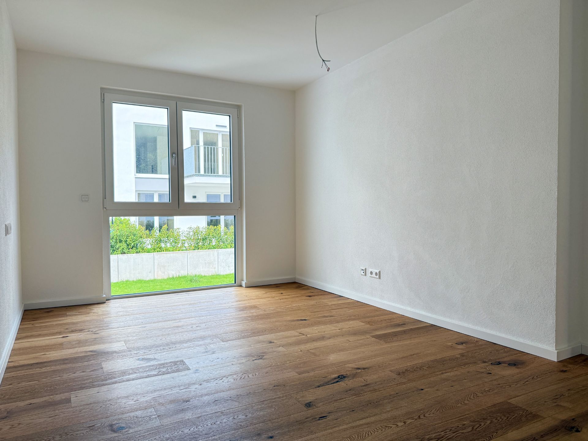 Leerer Raum mit Holzfußboden, weißen Wänden und großem Fenster mit Blick ins Grüne.