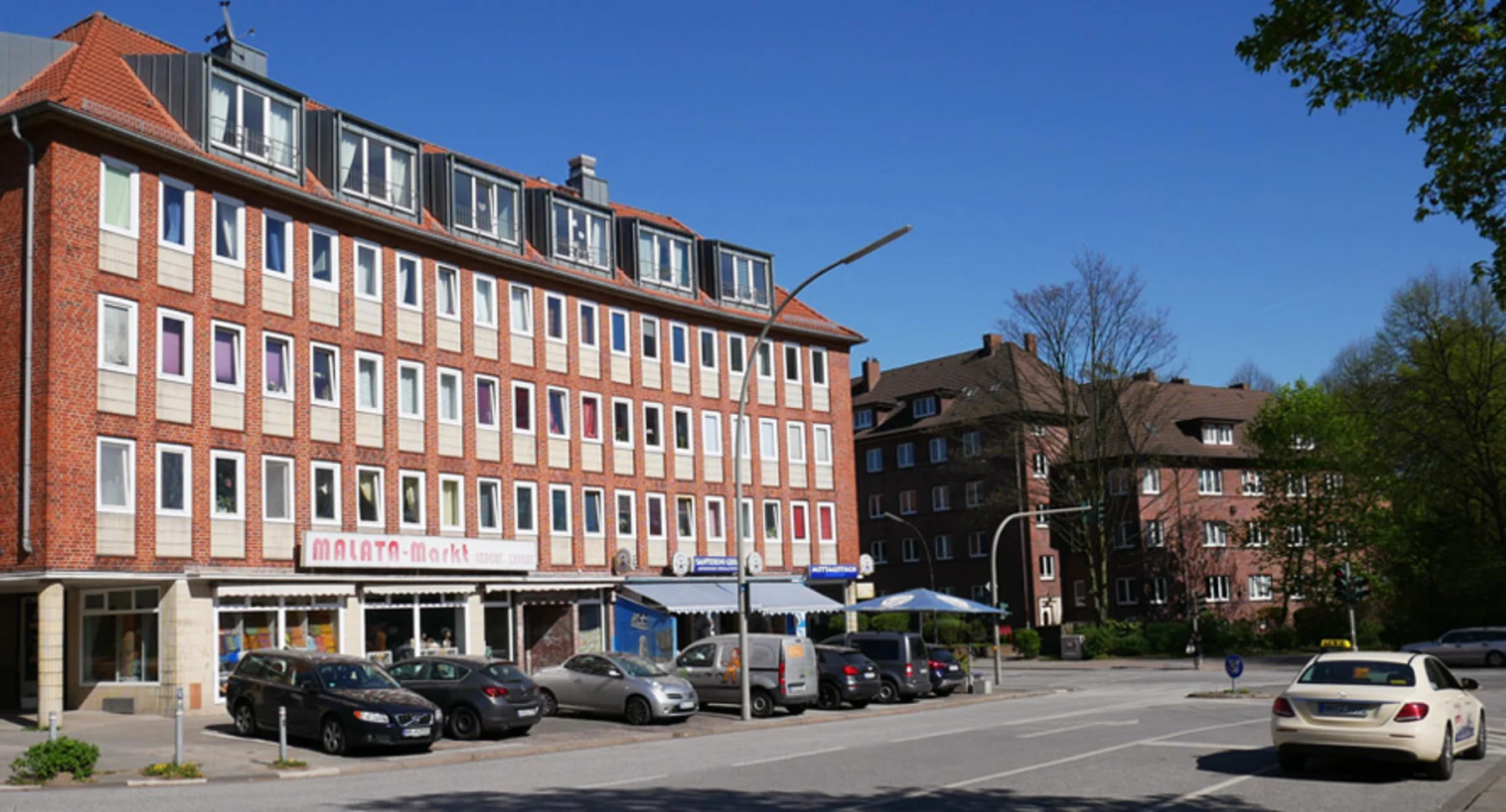 Mehrstöckiges Backsteingebäude mit Geschäften im Erdgeschoss und geparkten Autos davor.