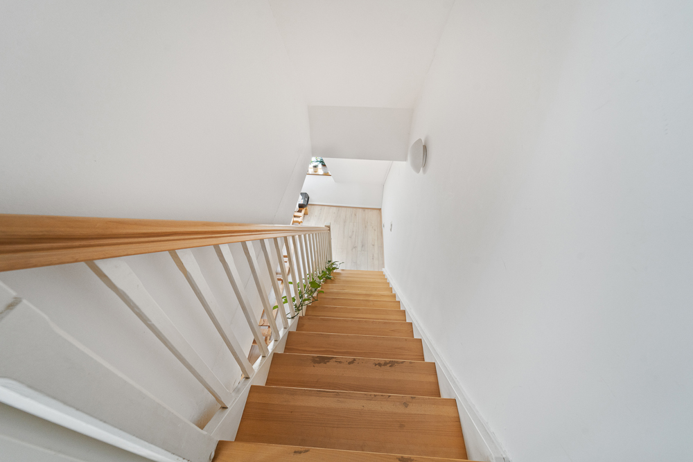 Hölzerne Treppe mit weißem Geländer in einem hellen Flur, Pflanze am Treppenrand.