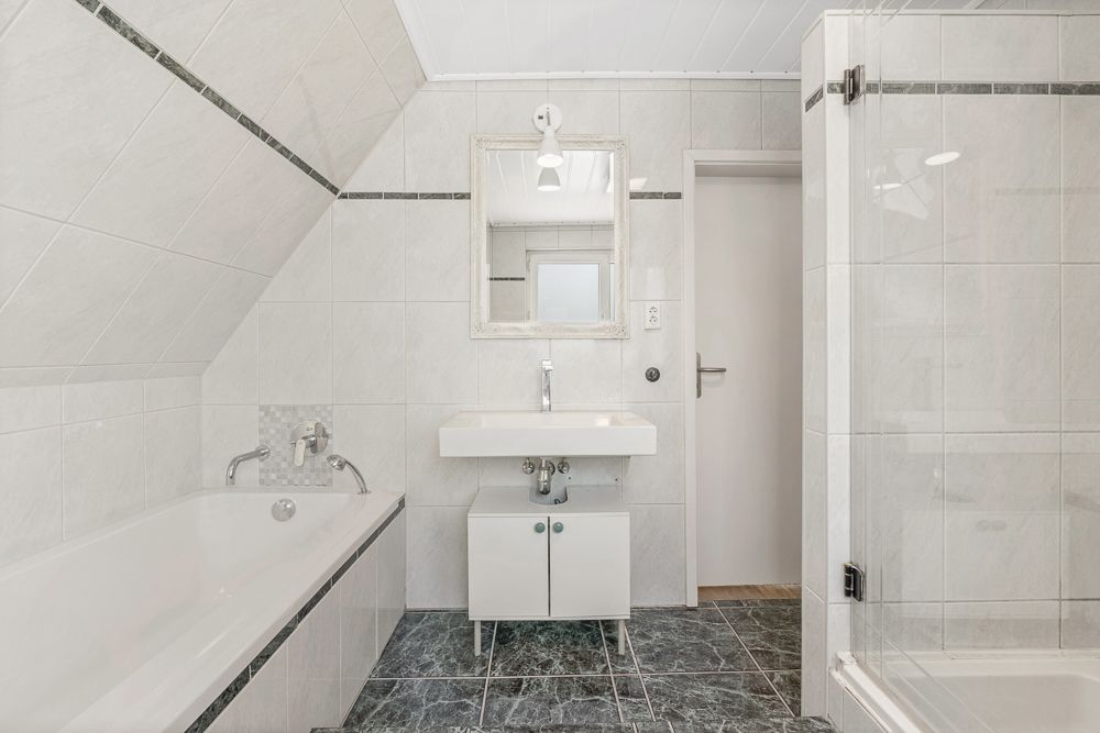 Helles Badezimmer mit Badewanne, Waschbecken, Spiegel und Dusche mit Glastür.