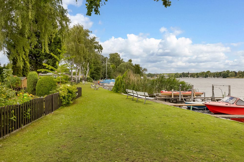 Grüne Wiese mit Bänken am Wasser, Boote am Steg und Bäume im Hintergrund.