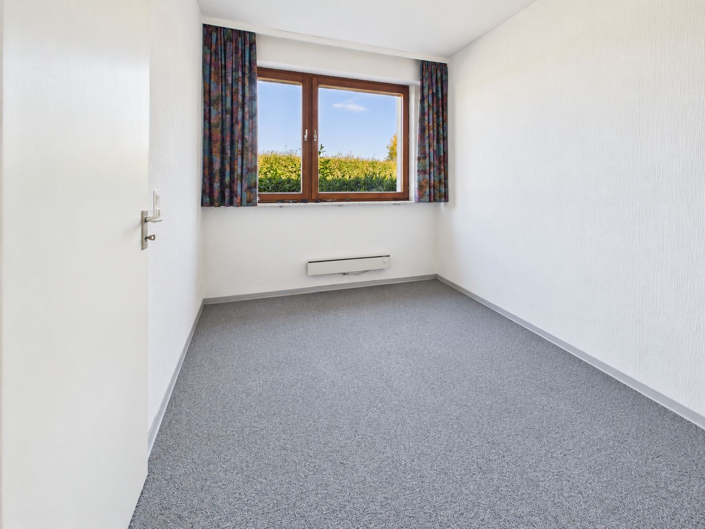 Leerer Raum mit grauem Teppich, weißem Wänden und Fenster mit bunten Vorhängen.