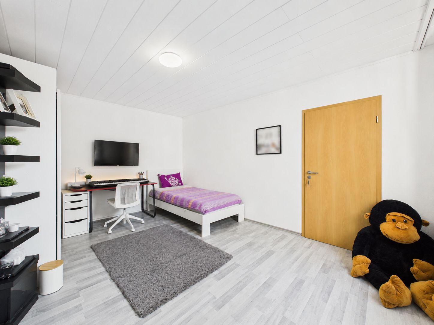 Helles Schlafzimmer mit Einzelbett, Schreibtisch, Fernseher und großem Plüschaffen auf dem Boden.