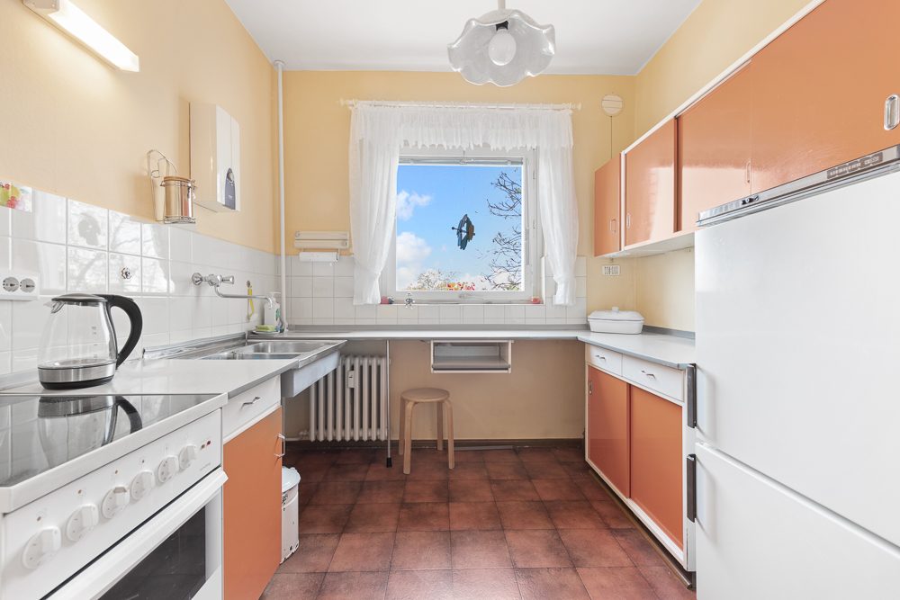 Helle Küche mit orangefarbenen Schränken, Herd, Kühlschrank und großem Fenster mit weißem Vorhang.
