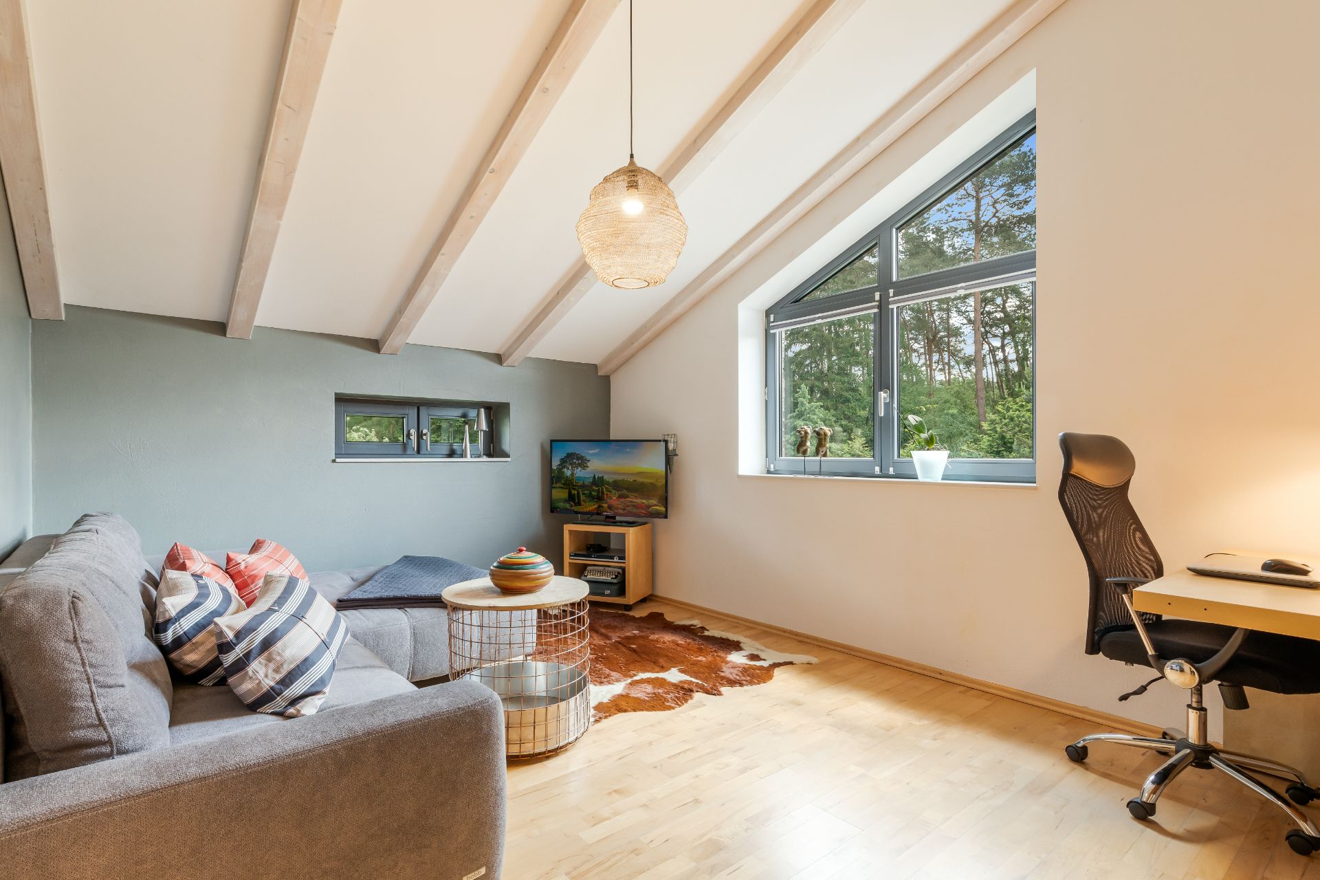 Helles Wohnzimmer mit Sofa, Schreibtisch, Fenster und Fernseher unter Dachschräge.
