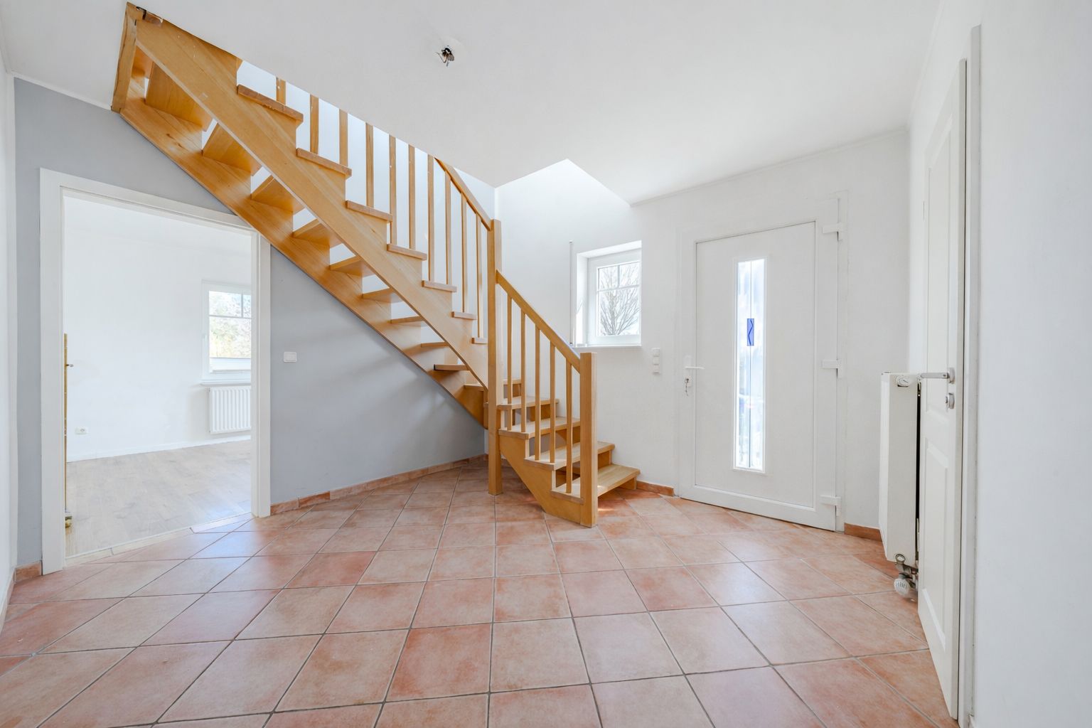 Heller Flur mit Fliesenboden und Holztreppe, Tür und Fenster, Zugang zu weiterem Raum.