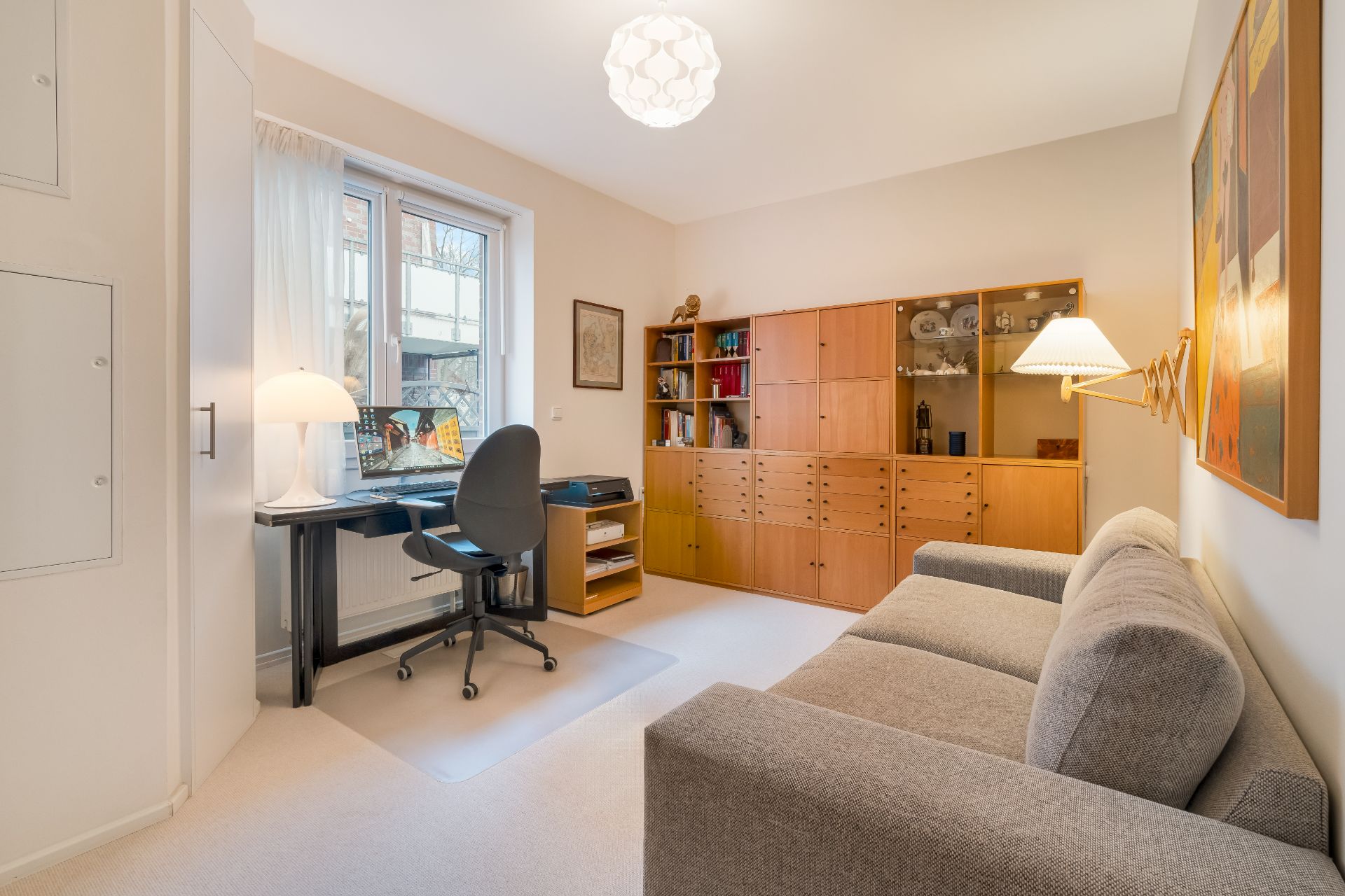 Helles Arbeitszimmer mit Schreibtisch, Regalwand, grauem Sofa und großem Fenster.