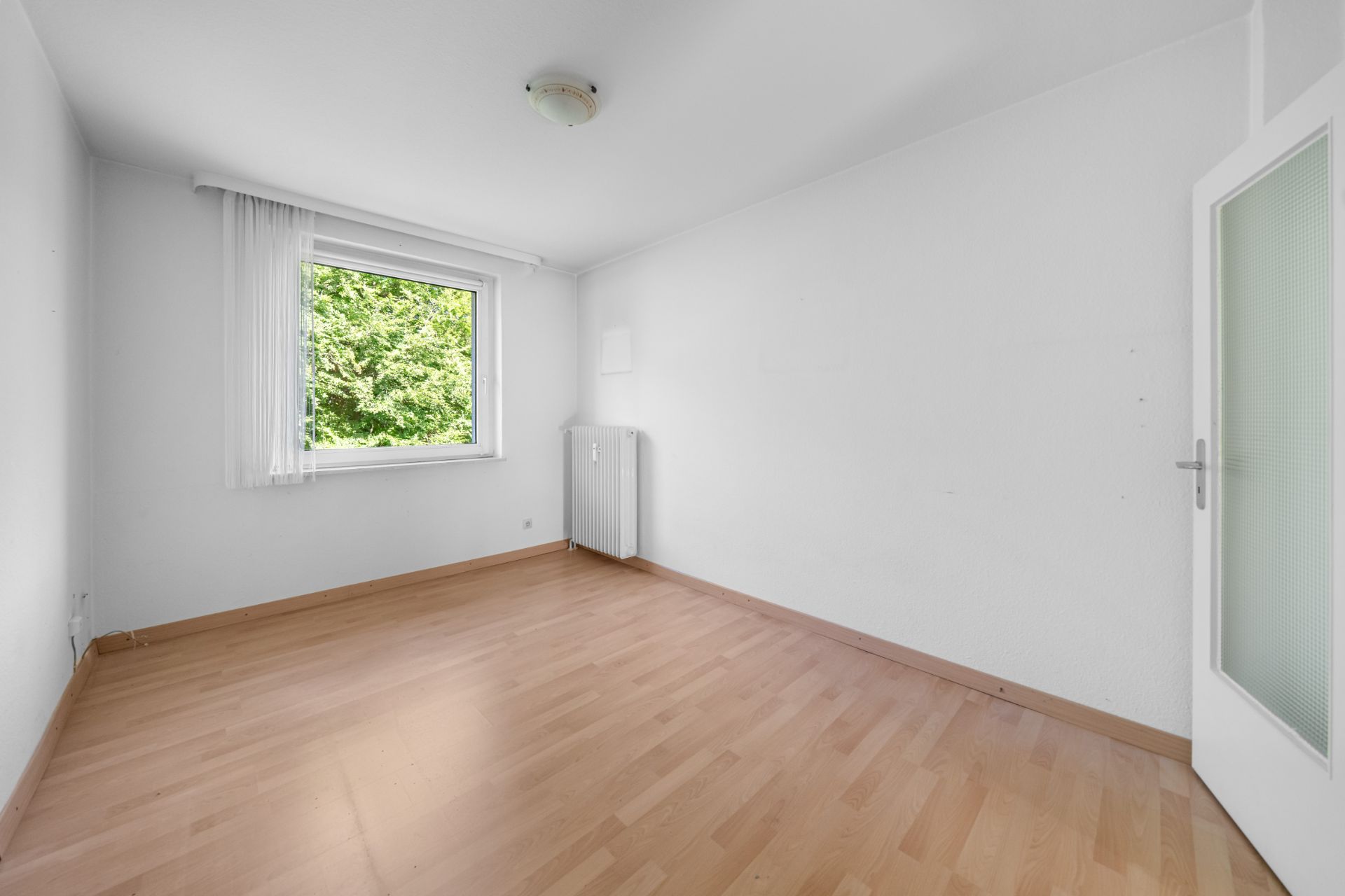 Leeres Zimmer mit Holzfußboden, weißer Wand, Fenster mit Vorhang und Heizkörper.