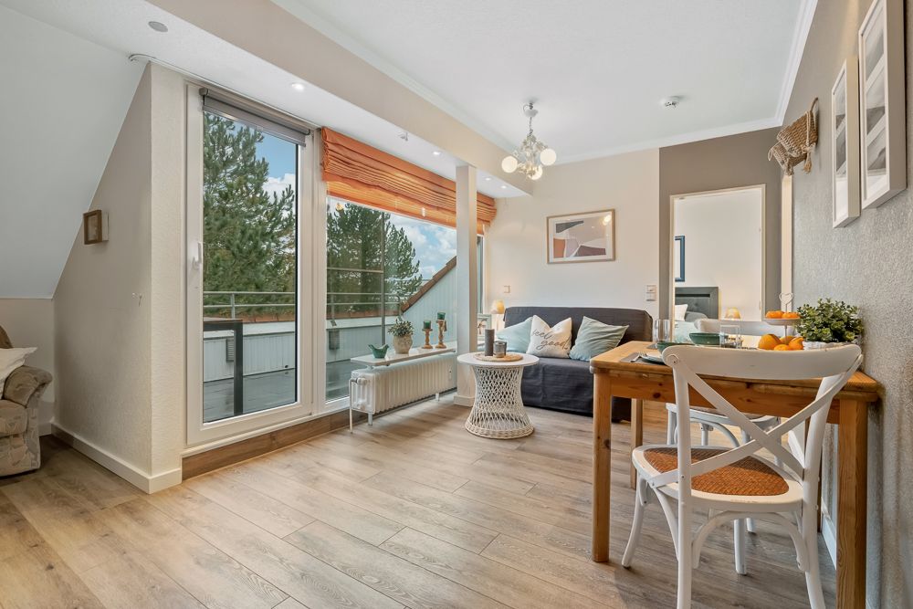 Helles Wohnzimmer mit Holzboden, Sofa, kleinem Esstisch und großen Fenstern mit Blick auf Bäume.