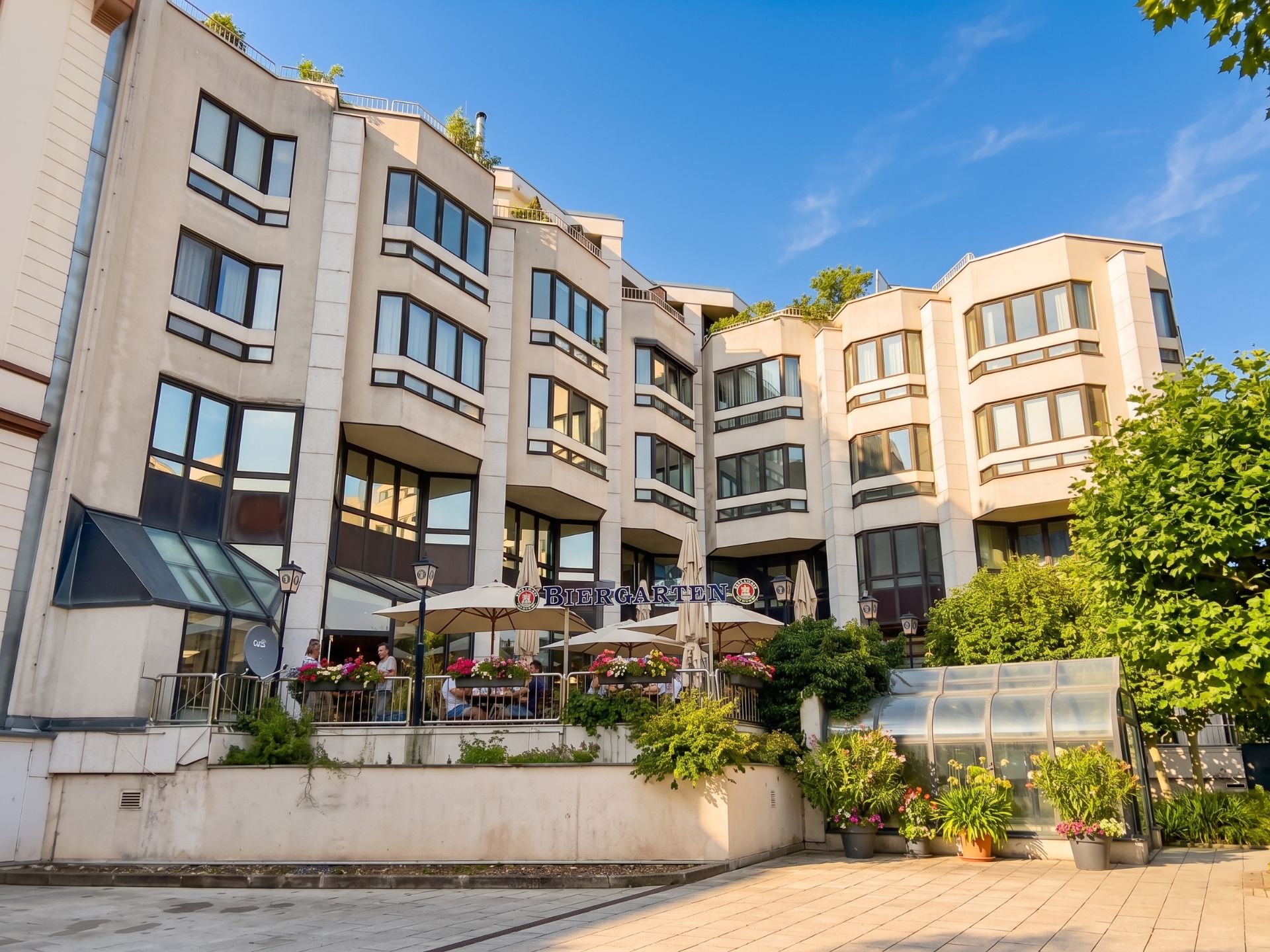 Mehrstöckiges Wohngebäude mit großen Fenstern und Terrasse mit Sonnenschirmen und Pflanzen.