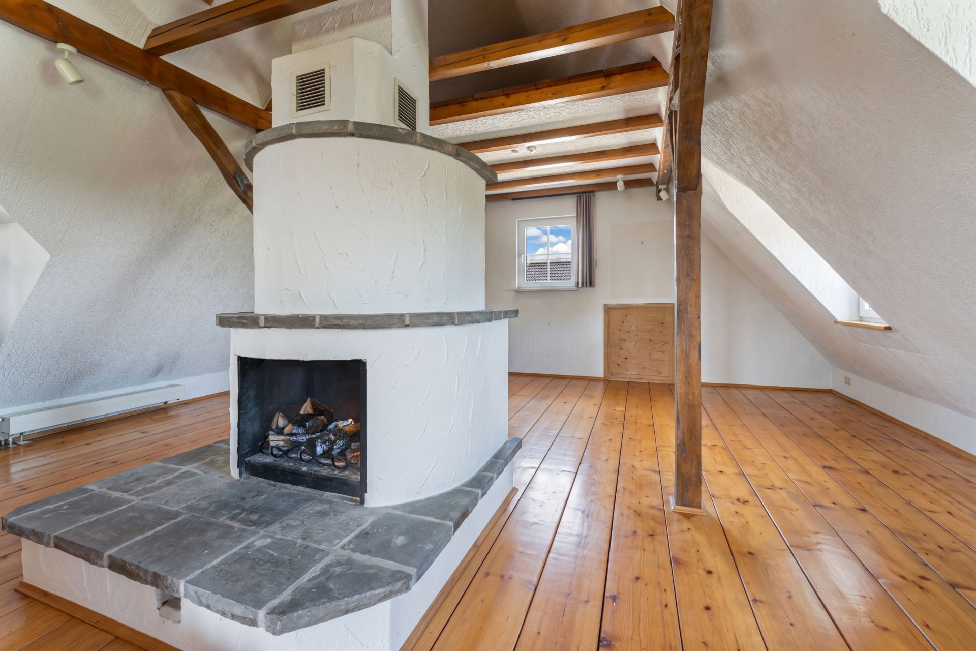Dachzimmer mit sichtbaren Holzbalken, Holzboden und rundem Kamin in der Mitte.