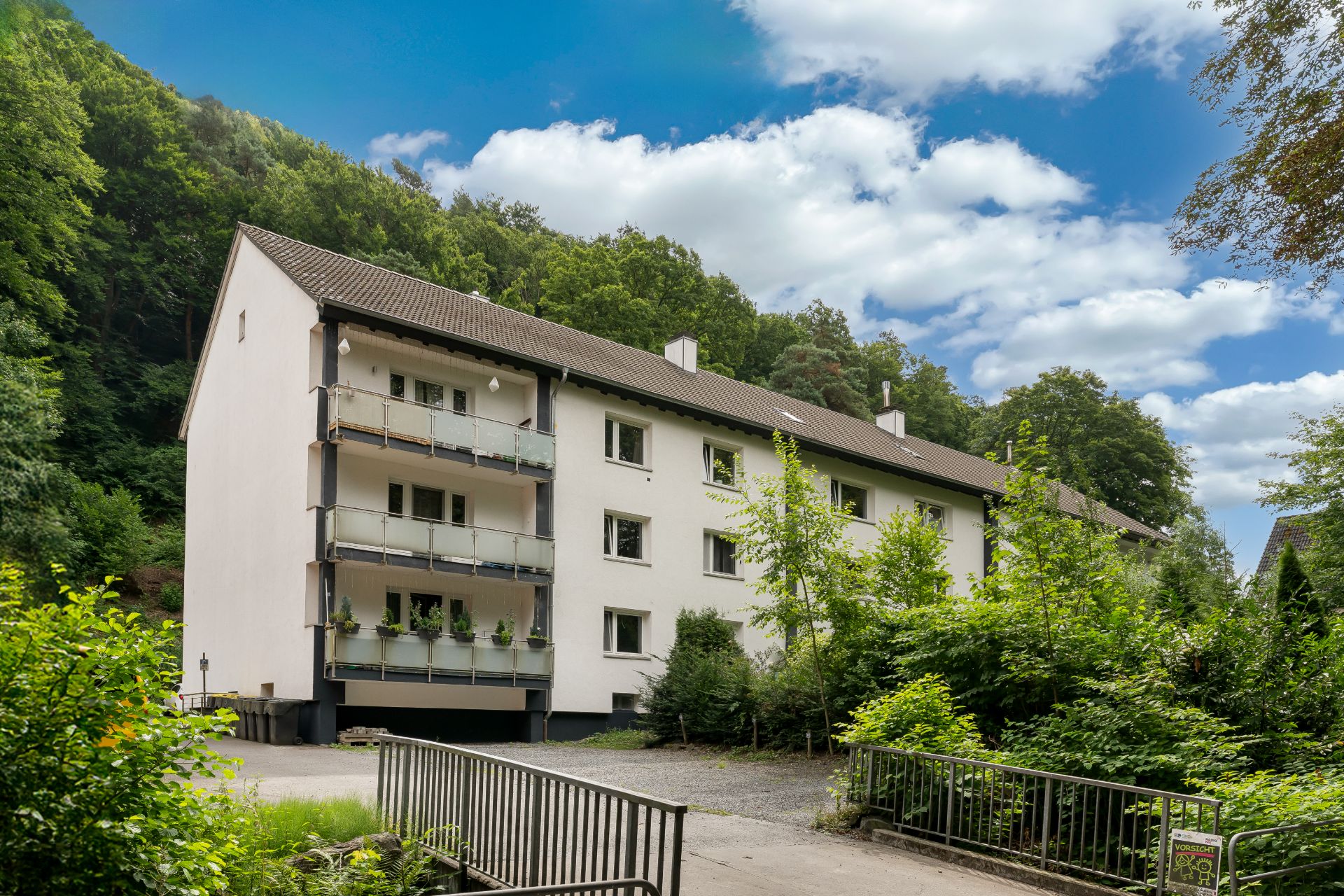 Mehrfamilienhaus mit Balkonen, umgeben von Bäumen und einer Einfahrt im Vordergrund.