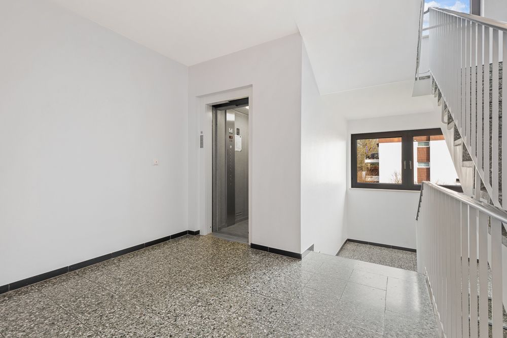 Heller Hausflur mit offenem Aufzug, Treppenhaus und Fenster mit Blick nach draußen.