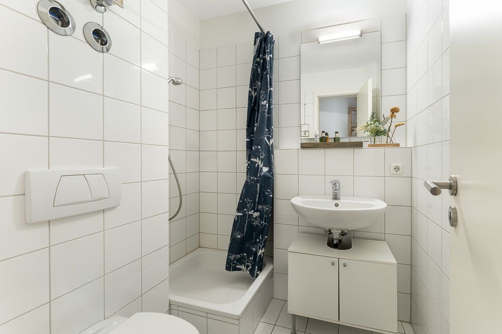 Kleines Badezimmer mit Dusche, Waschbecken, Spiegel und weiß gefliesten Wänden.