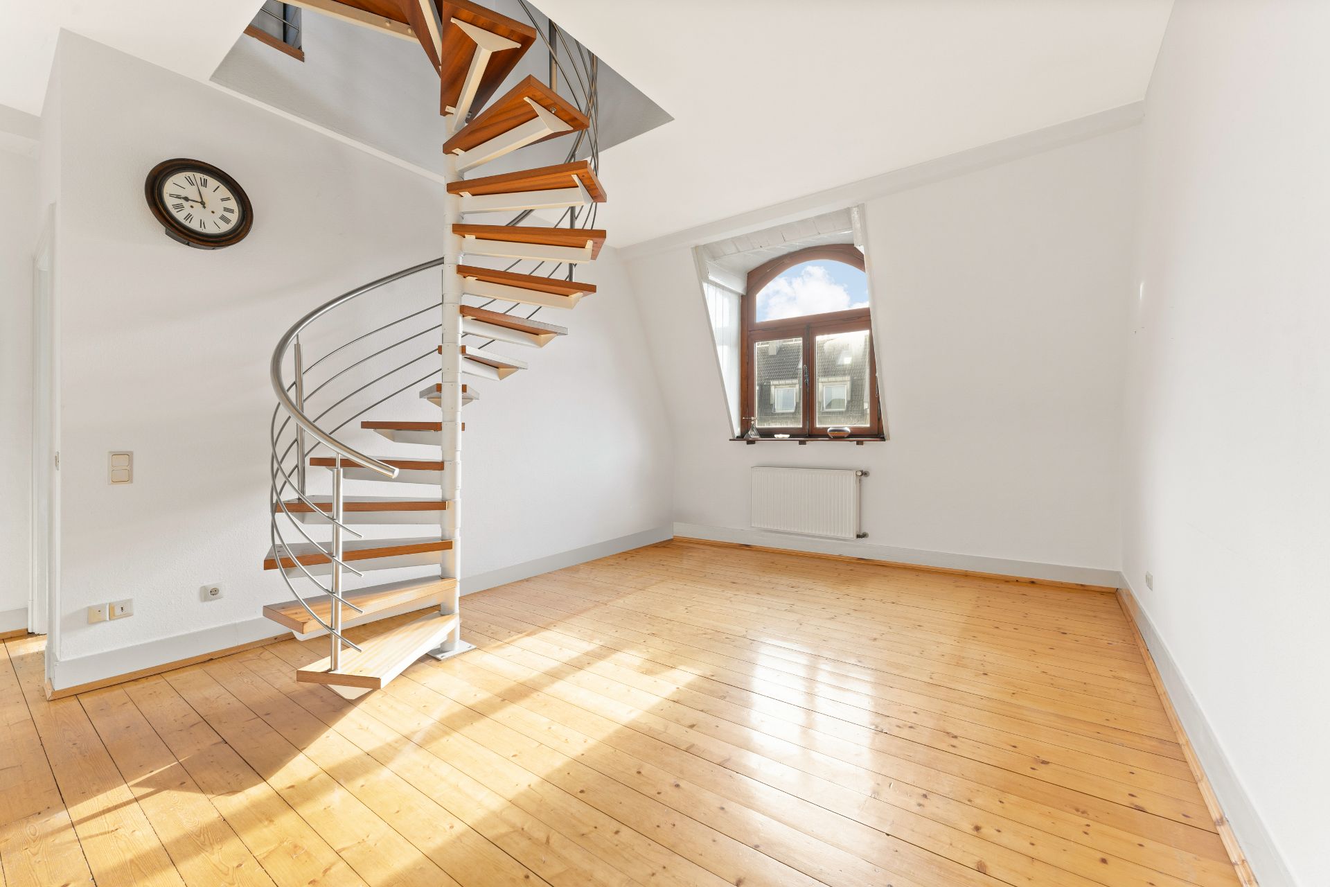 Helles Zimmer mit Holzfußboden, Wendeltreppe und Fenster mit Heizkörper darunter.