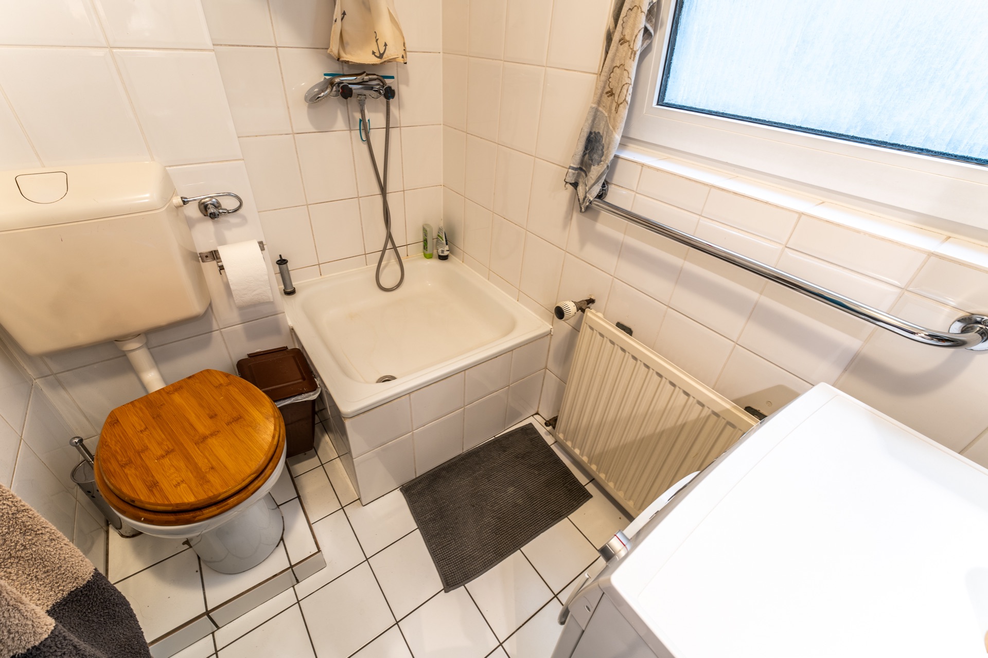 Kleines Badezimmer mit Toilette, Dusche, Waschmaschine, Heizung und einem Fenster.