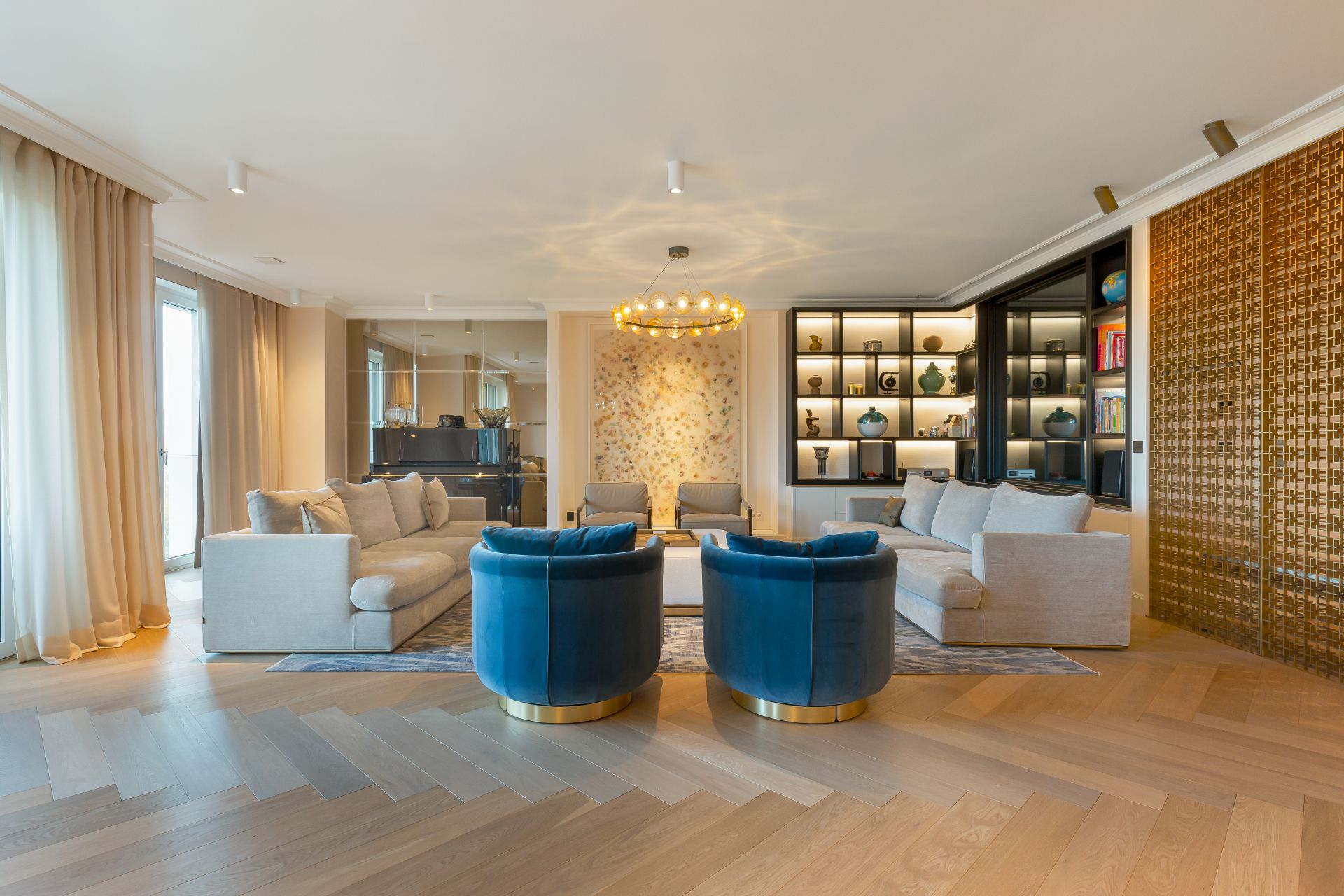 Modernes Wohnzimmer mit beigen Sofas, zwei blauen Sesseln und Regalwand mit Dekorationen.