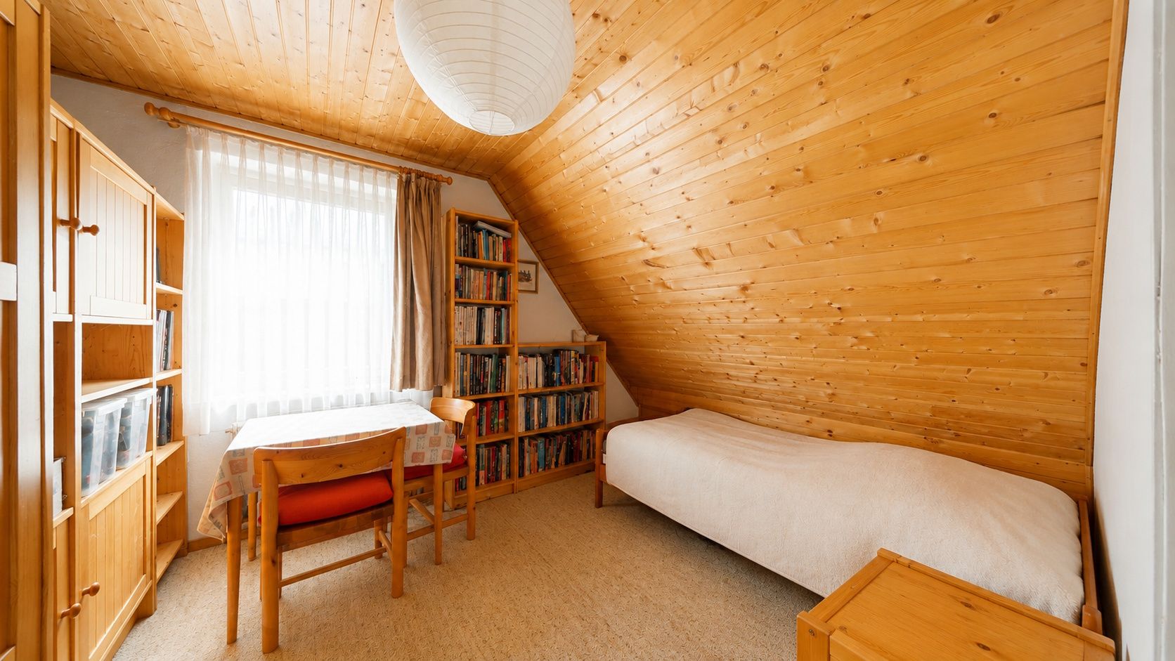 Kleines Zimmer mit Holzdecke, Einzelbett, Tisch, Bücherregalen und Fenster mit Vorhang.