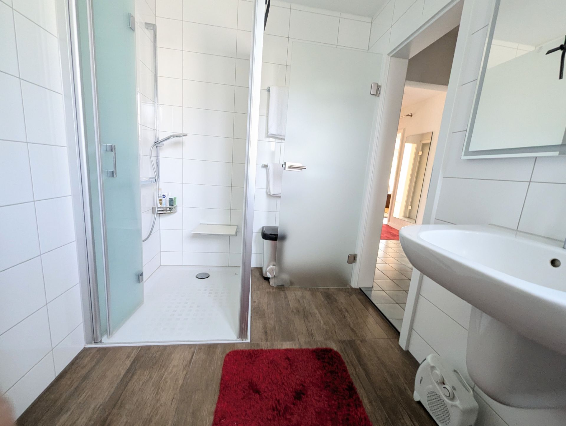 Helles Bad mit Dusche, Waschbecken, rotem Teppich und Fliesenboden.