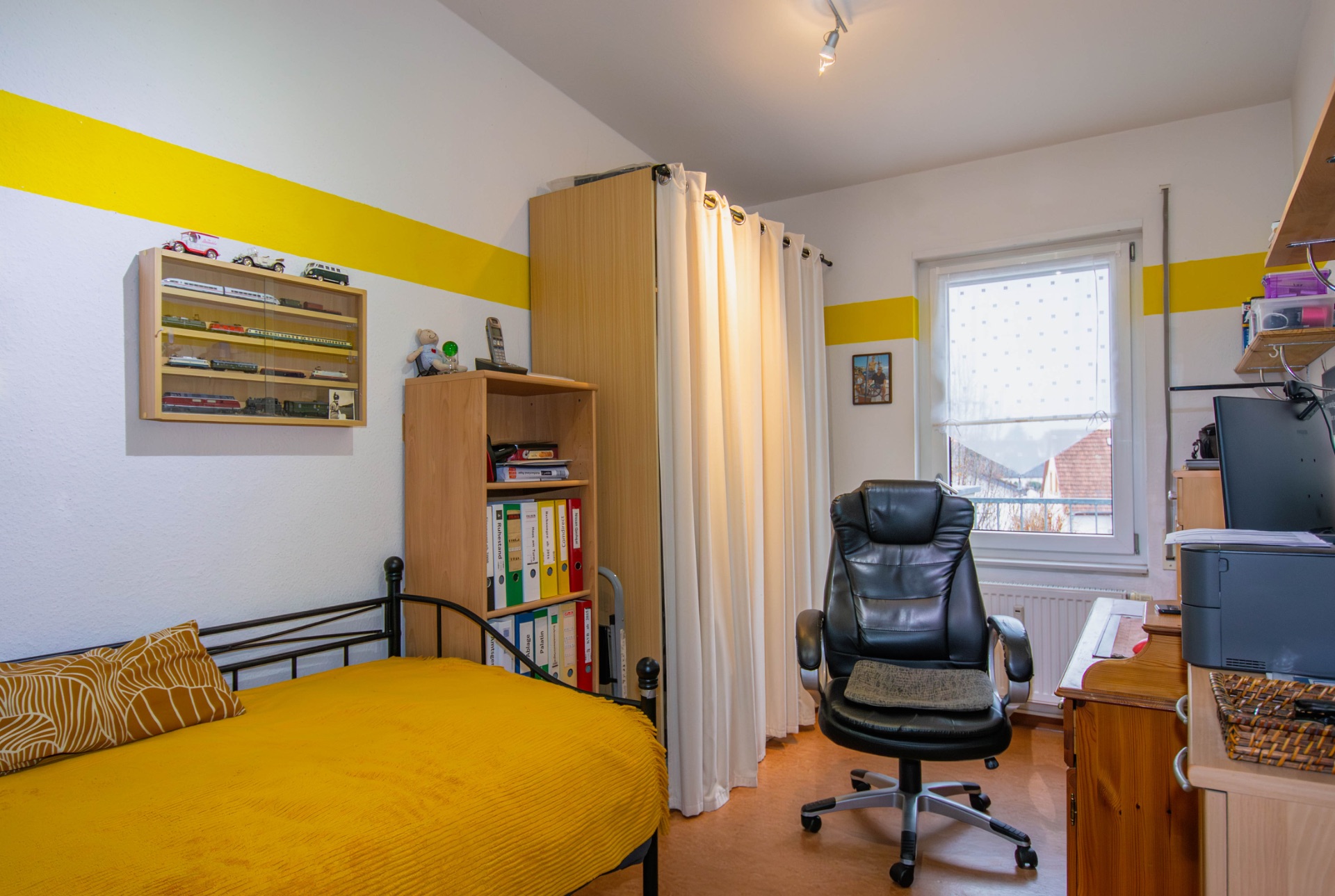 Kleines Zimmer mit Einzelbett, Schreibtisch, Bürostuhl und Regalen, gelber Akzentstreifen an der Wand.