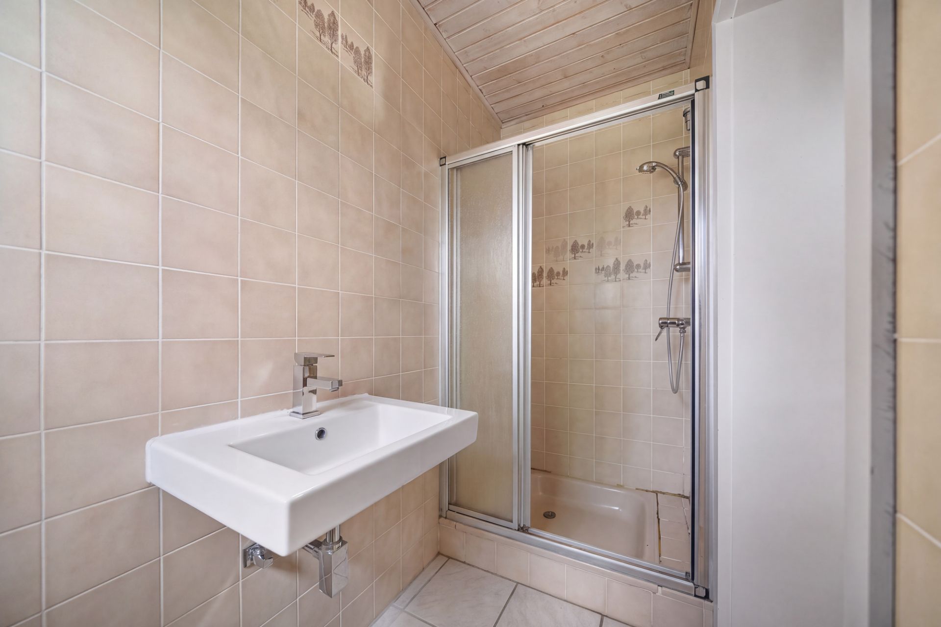 Kleines Badezimmer mit Waschbecken und Dusche mit Glastüren, beige Wandfliesen.